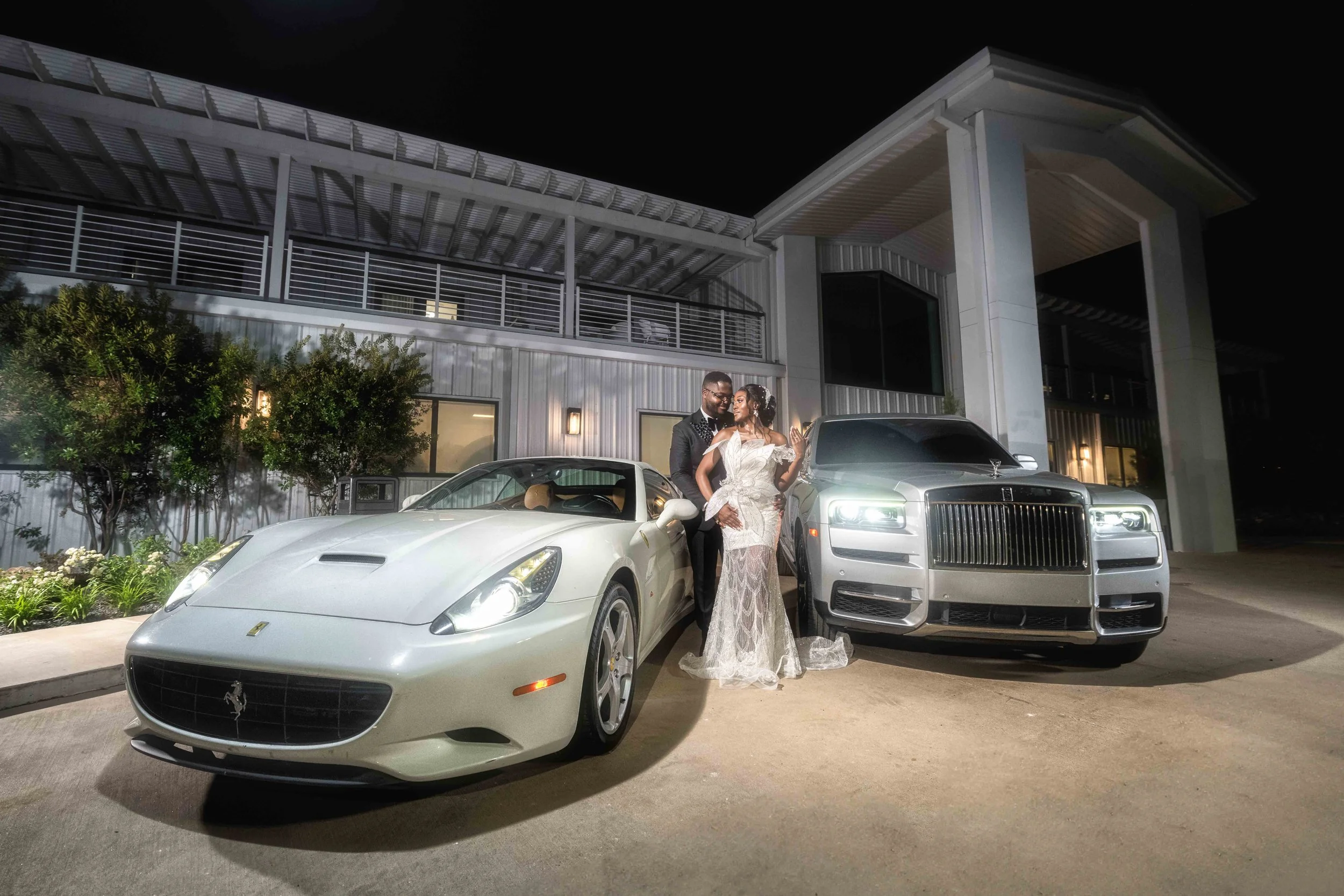 Couple dressed in wedding attire standing between a white Ferrari and a silver Rolls-Royce at night outside a modern building.