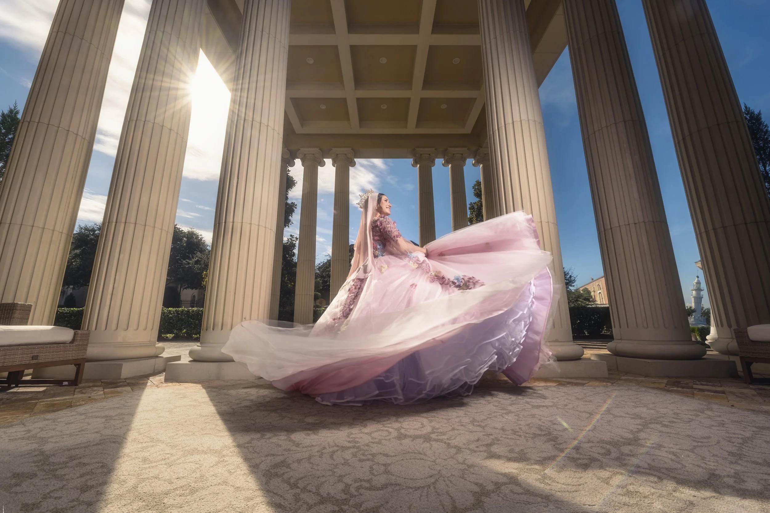 A woman in a pink wedding dress twirling under a Greek-style pavilion with tall columns, sunlight shining through, and blue sky in the background.