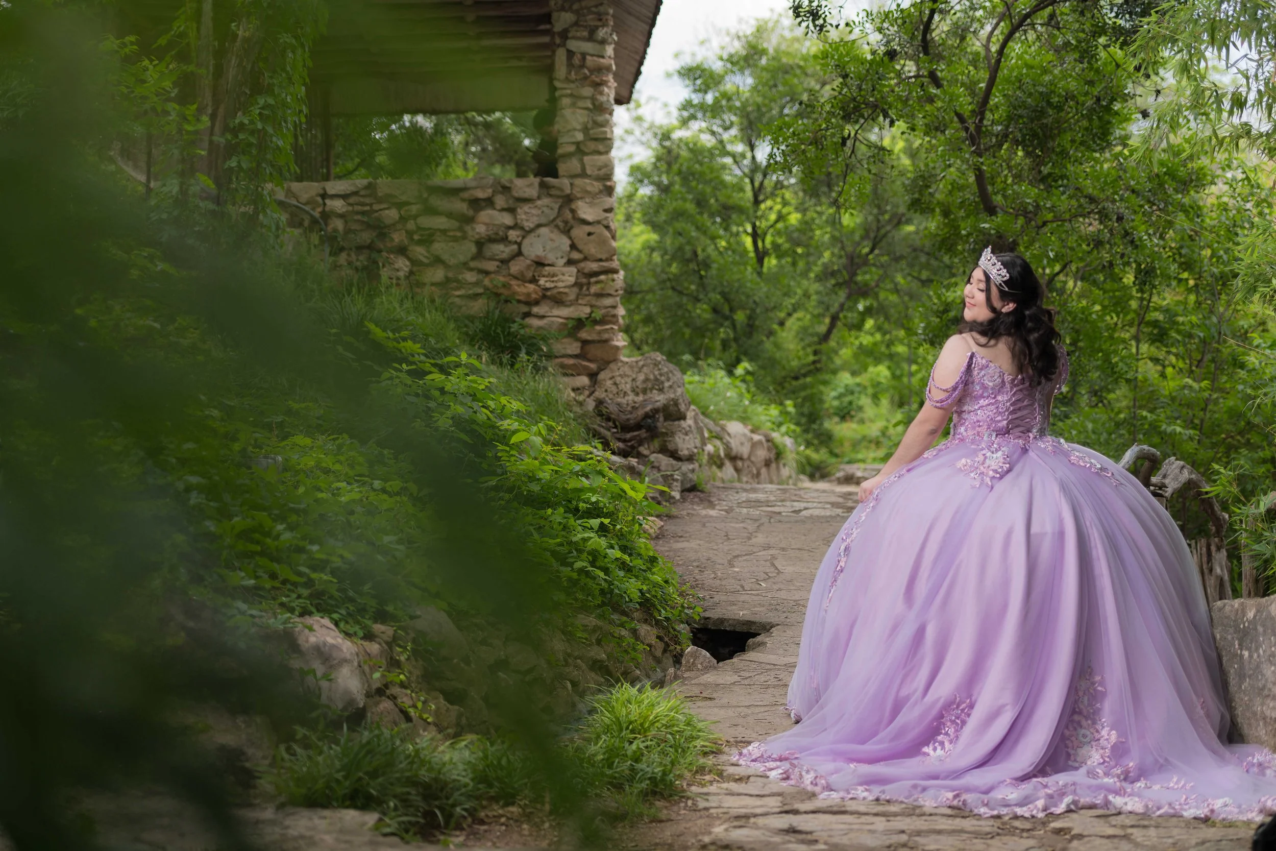 A woman in a lavender ballerina-style gown and tiara sitting outdoors on a stone bench, surrounded by green trees and foliage. She looks content, with a stone building and pathway nearby.