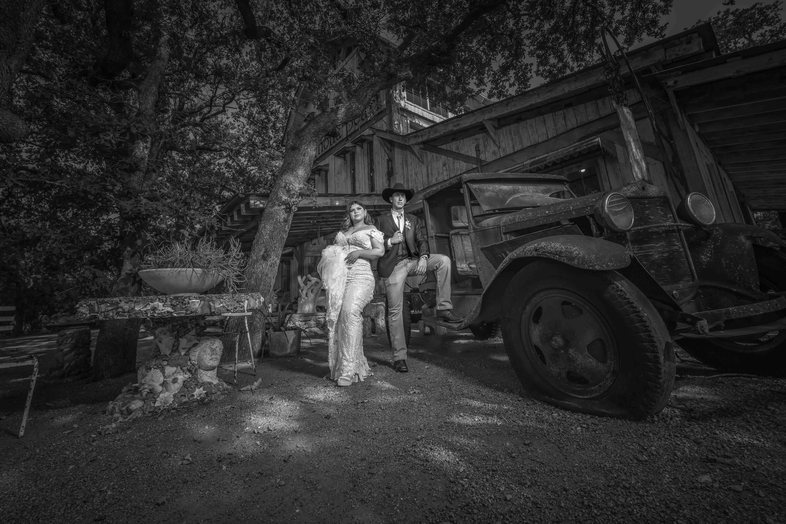 A black and white photo of a man and woman dressed in vintage clothing standing next to an old truck, with a wooden building and trees in the background.
