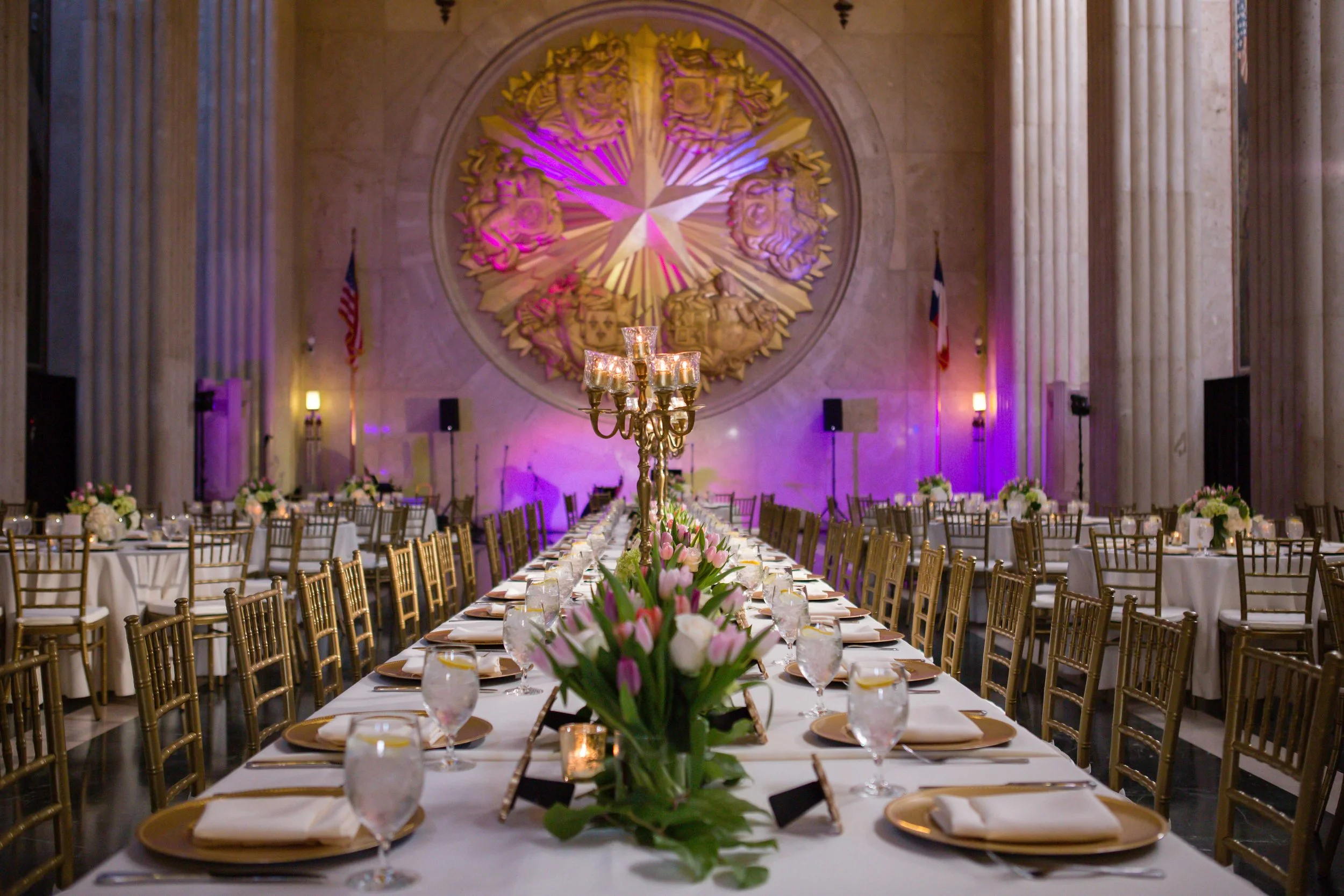 Elegant banquet hall set for a formal event with a long white table, floral centerpieces, gold chairs, and purple lighting, with large sculpture and American flags in the background.
