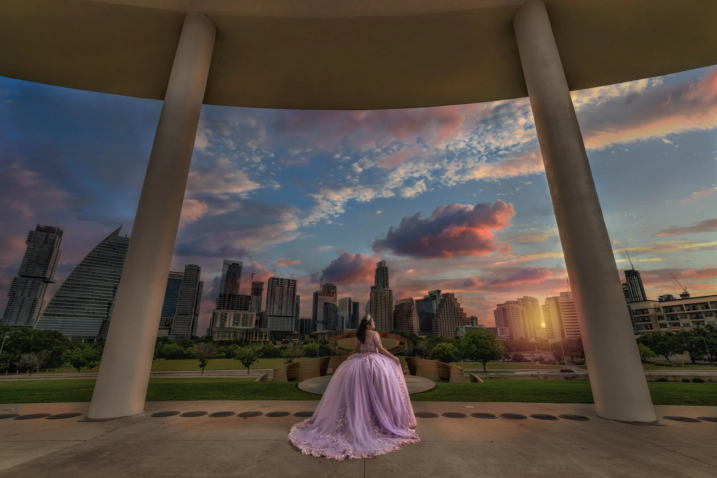 A woman in a lavender ball gown sitting on a circular bench at a park with a city skyline at sunset.