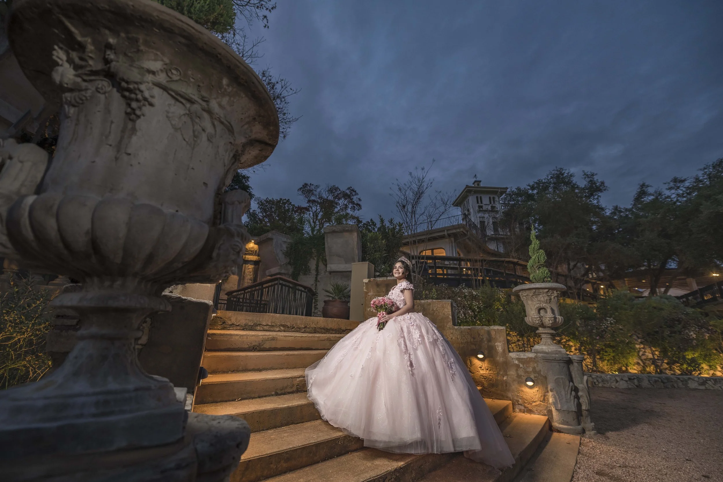 A woman in a pink wedding dress with floral embellishments, standing on stairs at dusk, holding pink flowers, and smiling, with decorative stone urns and trees in the background.