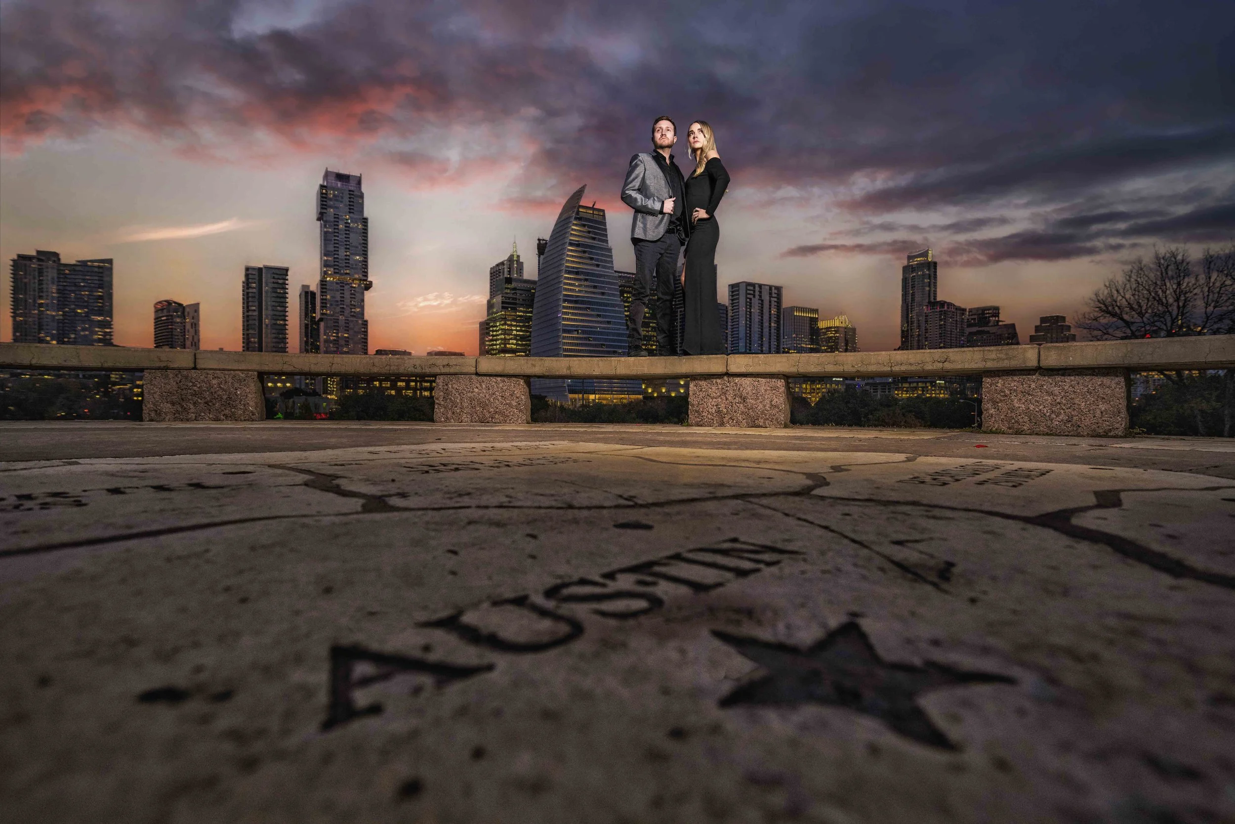 A man and woman dressed in formal attire standing on a stone ledge in front of a city skyline at dusk, with a partly cloudy sky and tall buildings in the background.