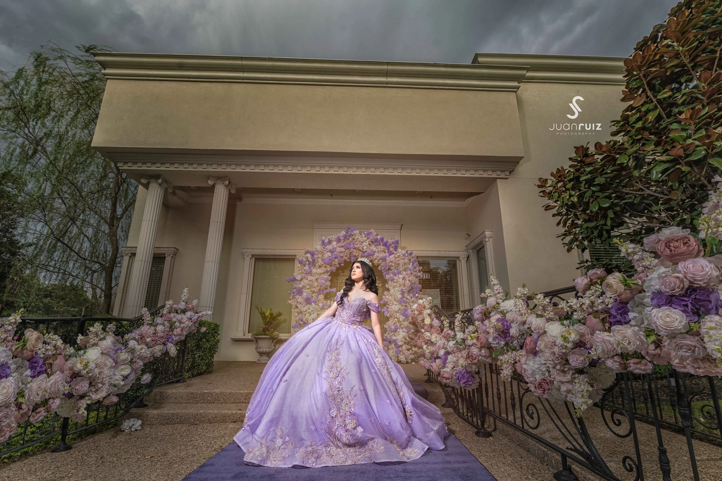 A woman in a lavender ball gown standing at the bottom of a floral staircase in front of a mansion.