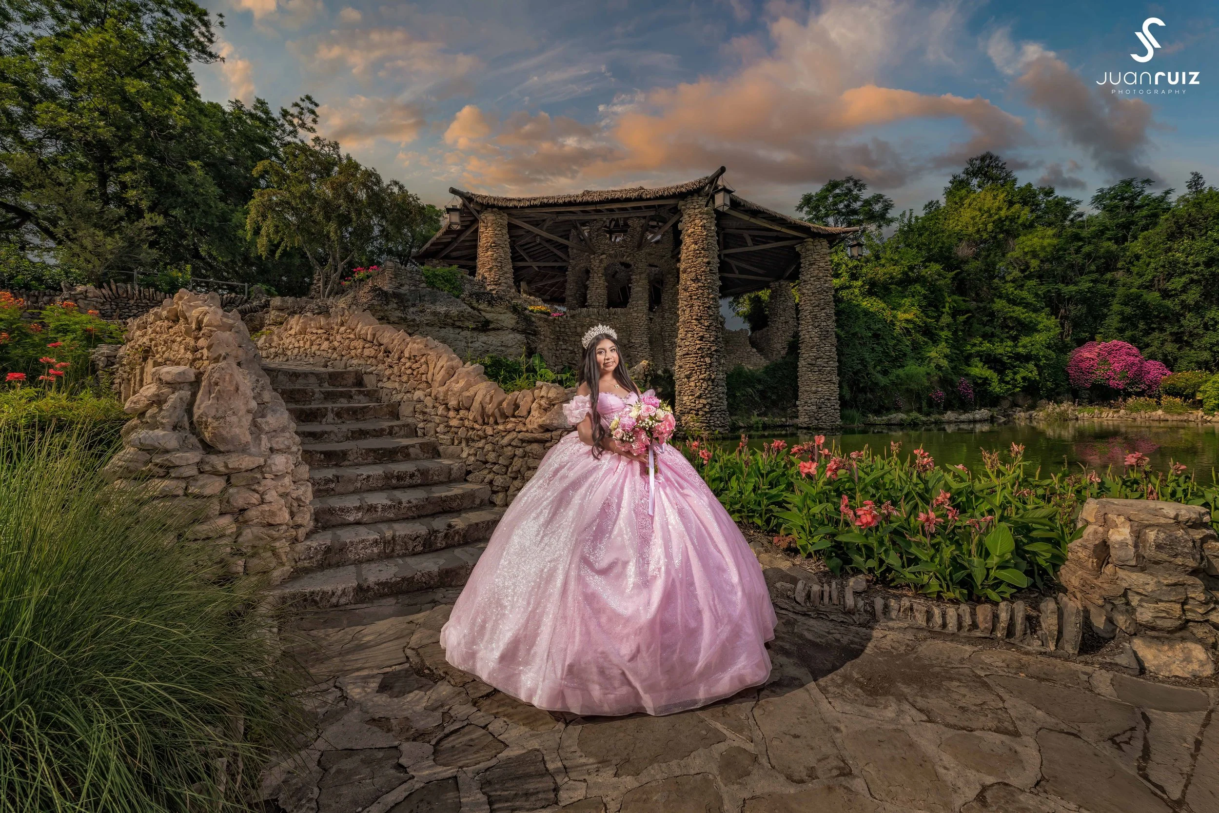 A young woman in a pink princess dress holding a bouquet of flowers standing on stone steps near a pond with colorful flowers and greenery, with a rustic stone pavilion in the background, during sunset.
