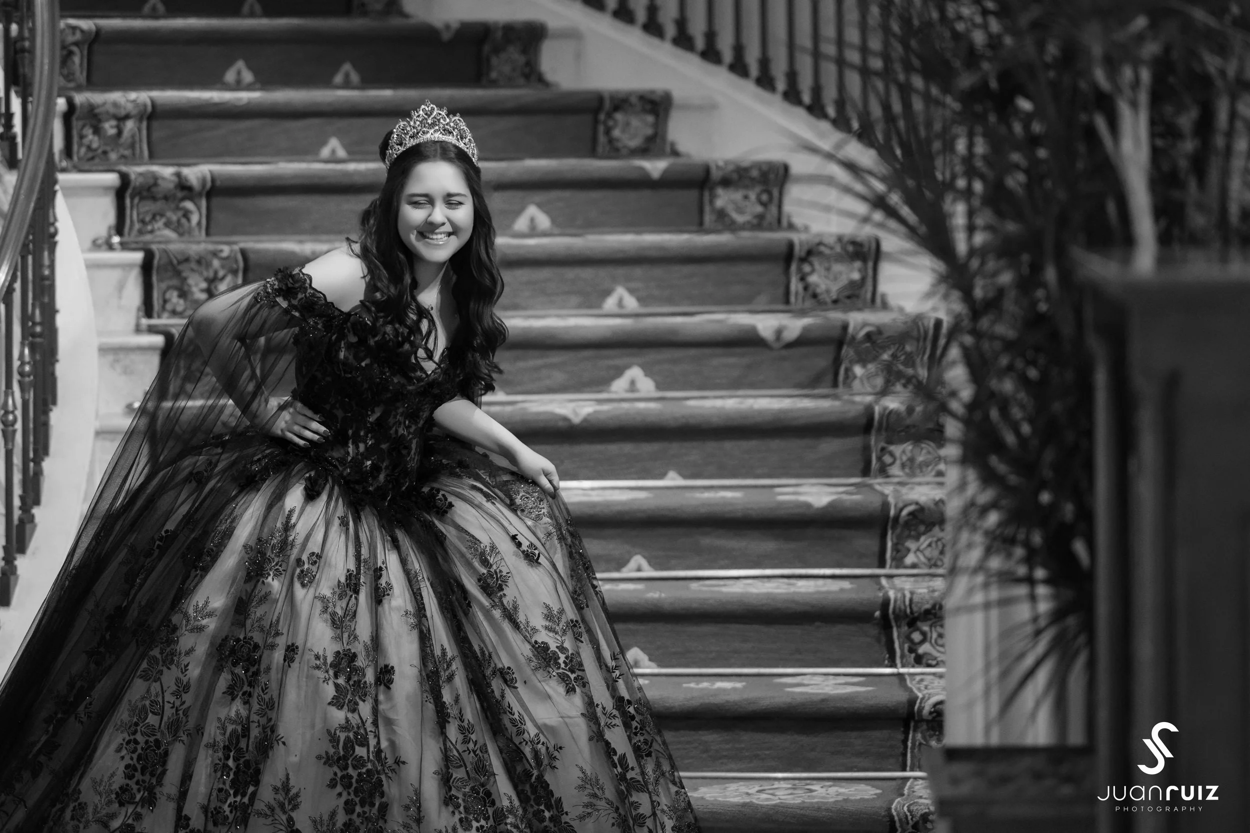 A young woman in a formal gown and tiara, smiling and sitting on a staircase, black and white photo.