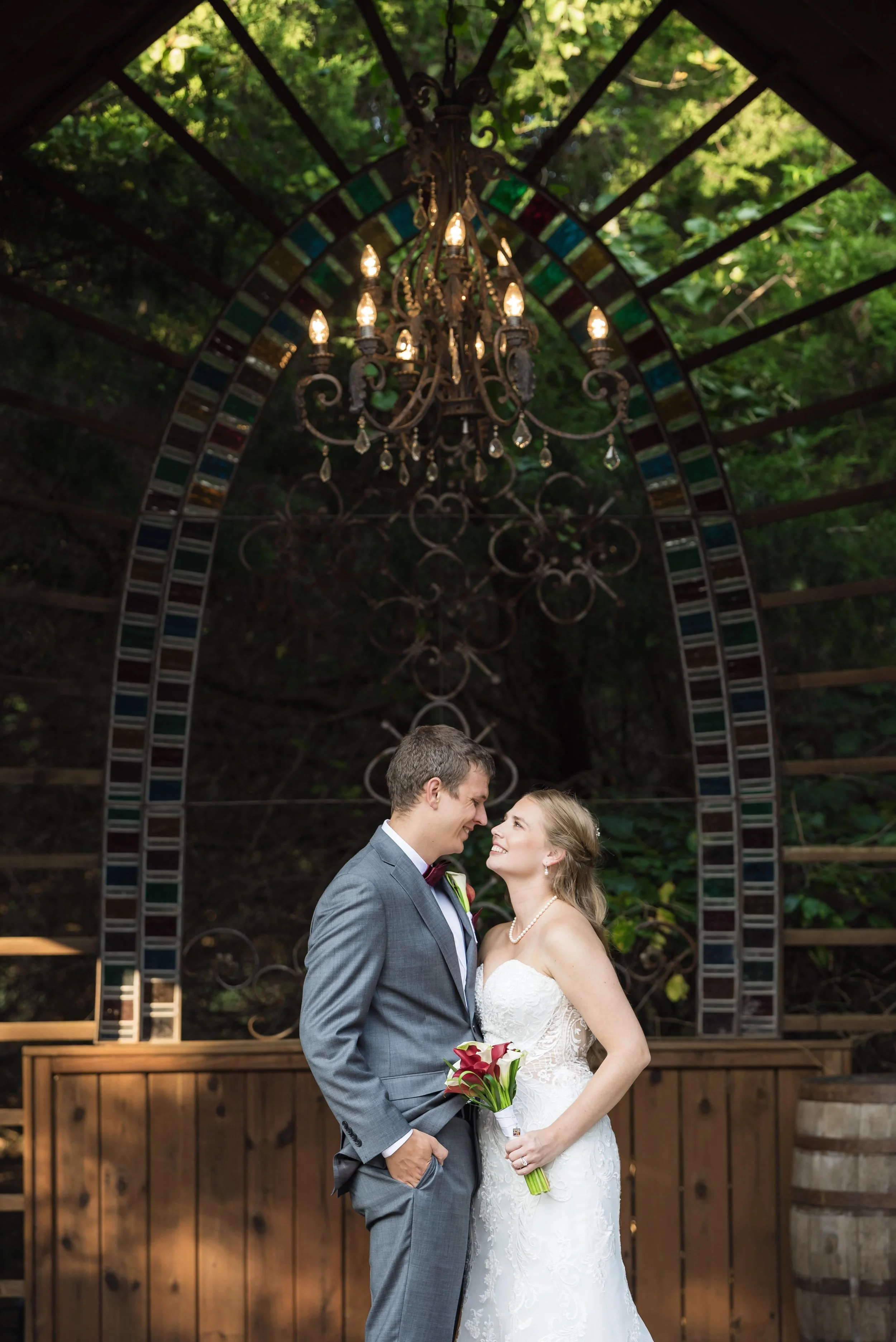 A bride and groom standing close together, smiling, in front of an ornate chandelier hanging under a wooden gazebo, surrounded by greenery.