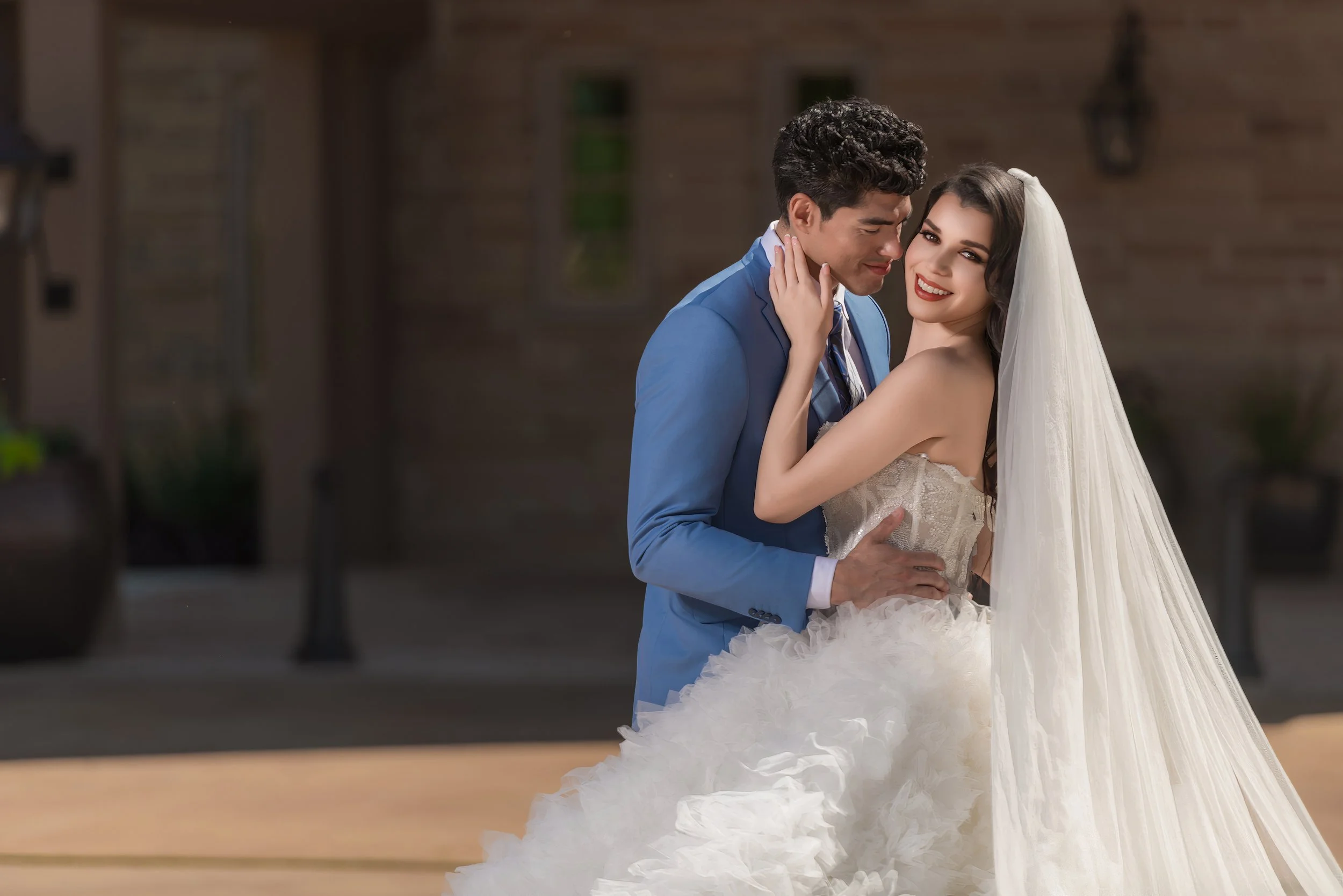 A bride and groom embracing outdoors, smiling, with the bride wearing a wedding dress and veil, and the groom in a blue suit.