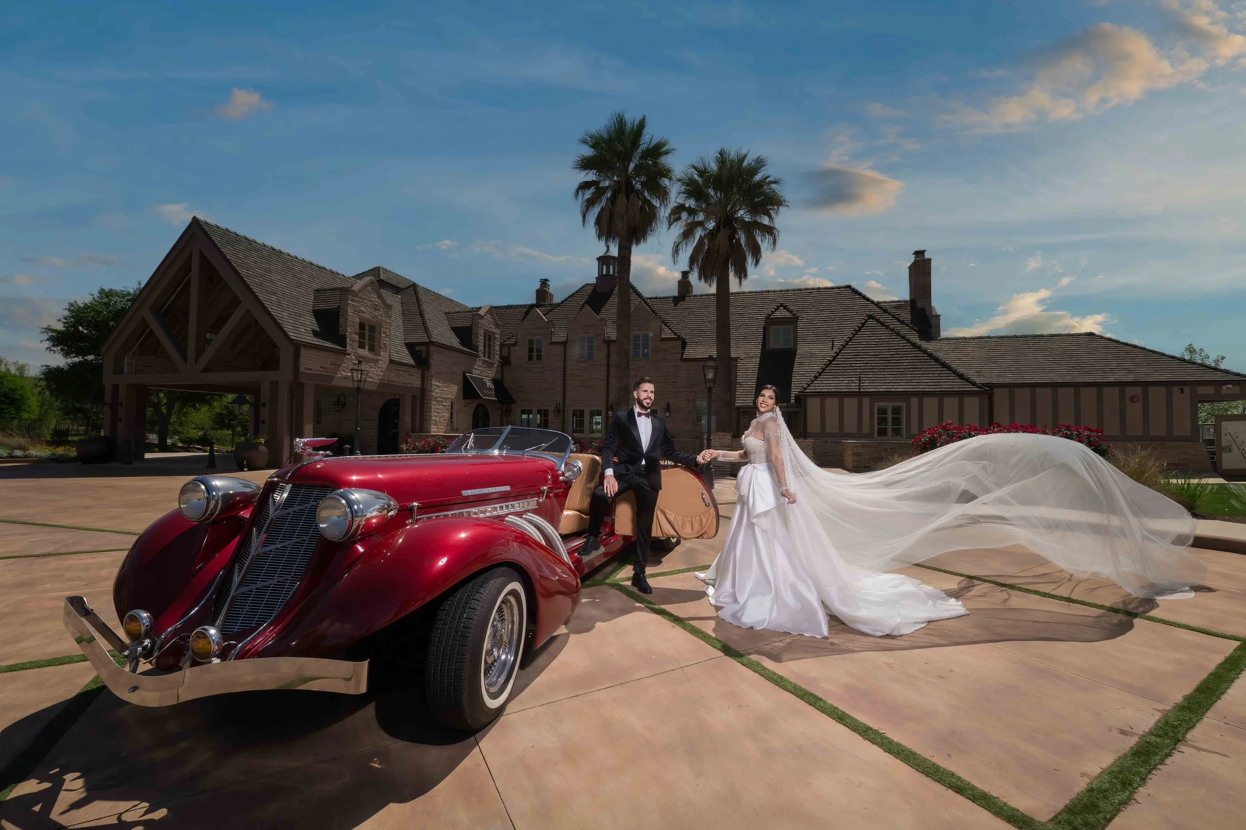 A bride and groom holding hands beside a vintage red convertible car in front of a large house with palm trees, on a sunny day.