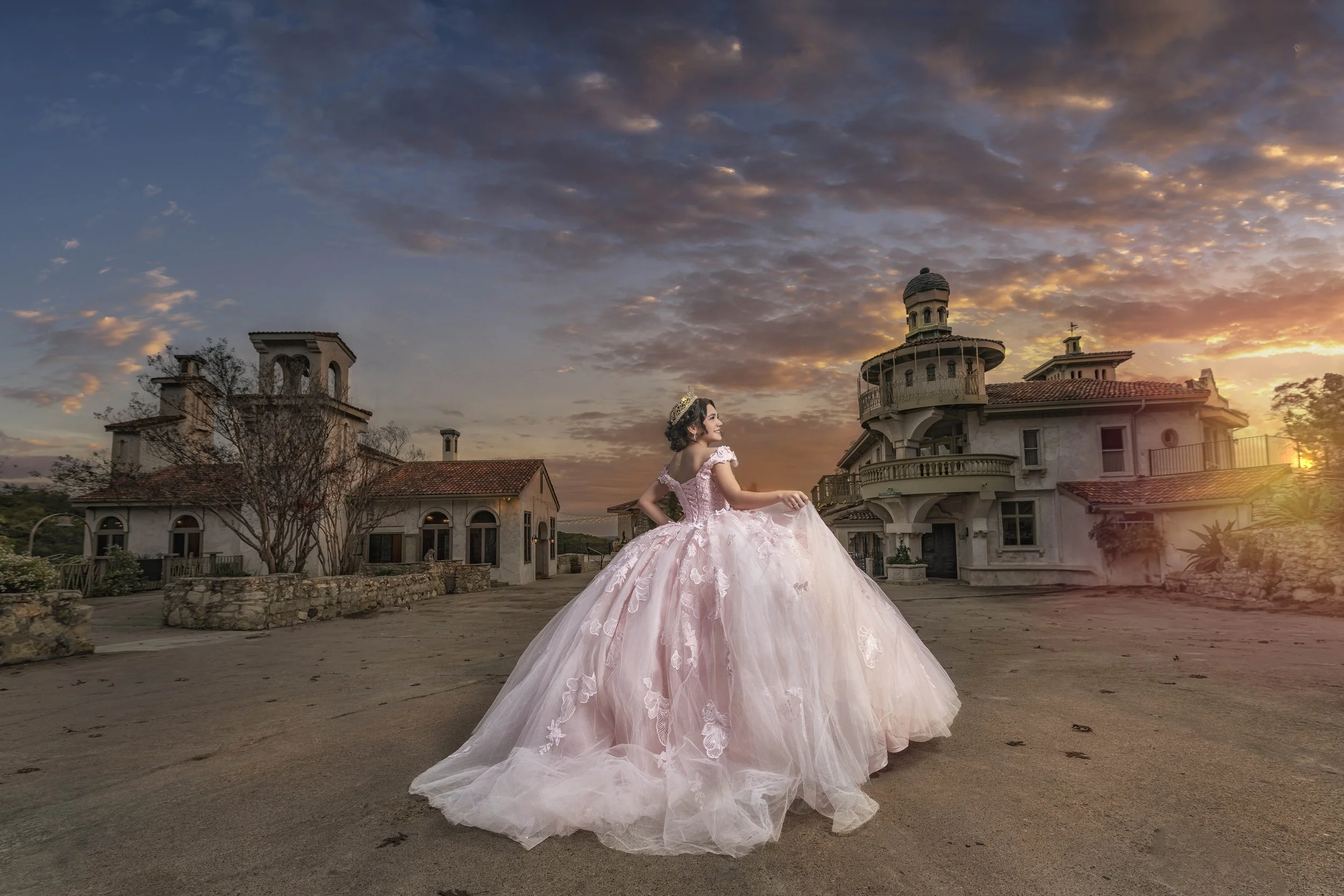 A woman in a pink ball gown and tiara standing on a dirt driveway in front of large, old-style buildings during sunset.