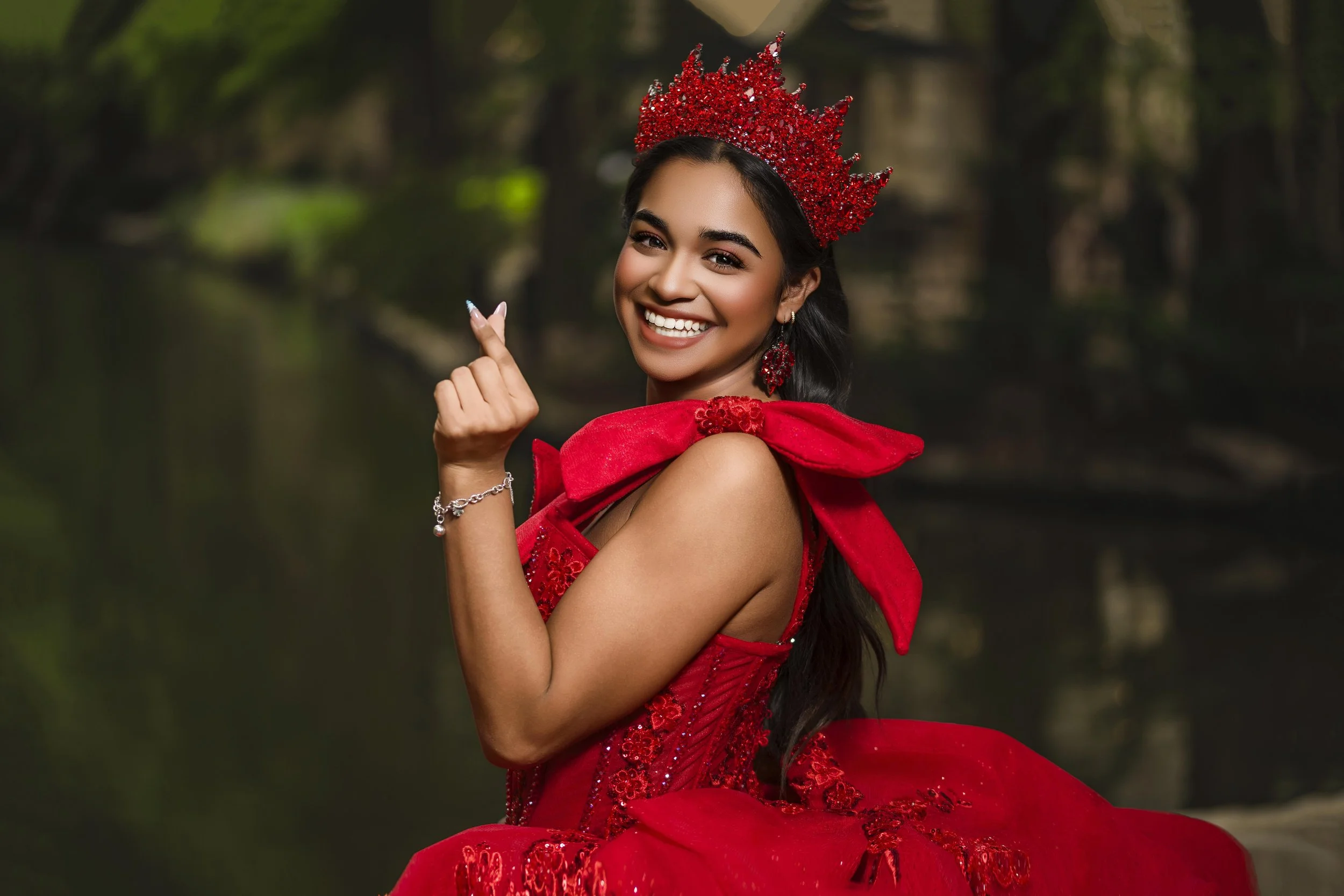 Young woman with long dark hair wearing a pink off-the-shoulder gown, tiara, earrings, and necklace, smiling and standing near a wooden wall.