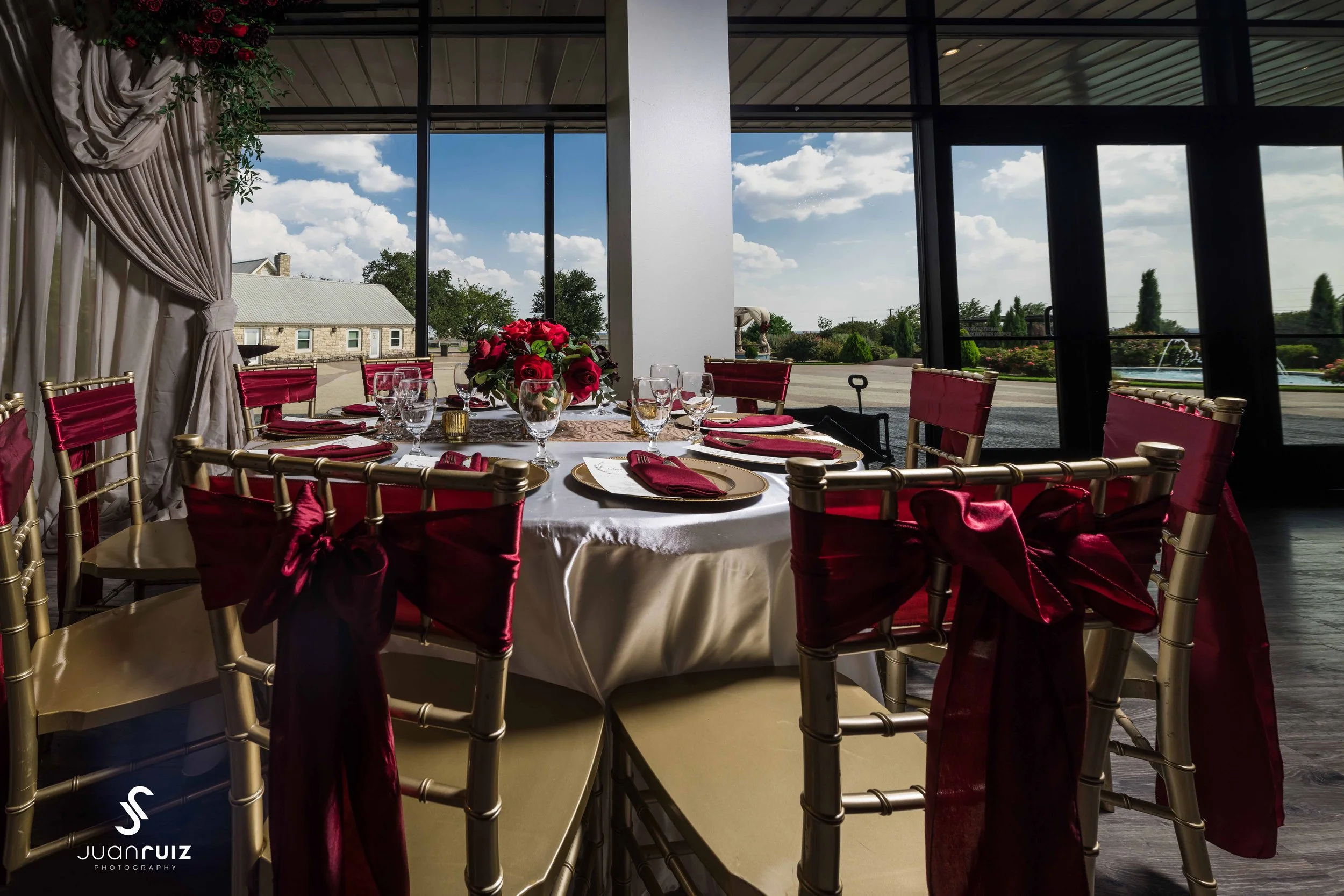 Elegant round dining table set with white tablecloth, red napkins, gold plates, glassware, and a floral centerpiece with red and pink flowers. The table is surrounded by gold chairs with red sashes, in a room with large glass windows showing a sunny 