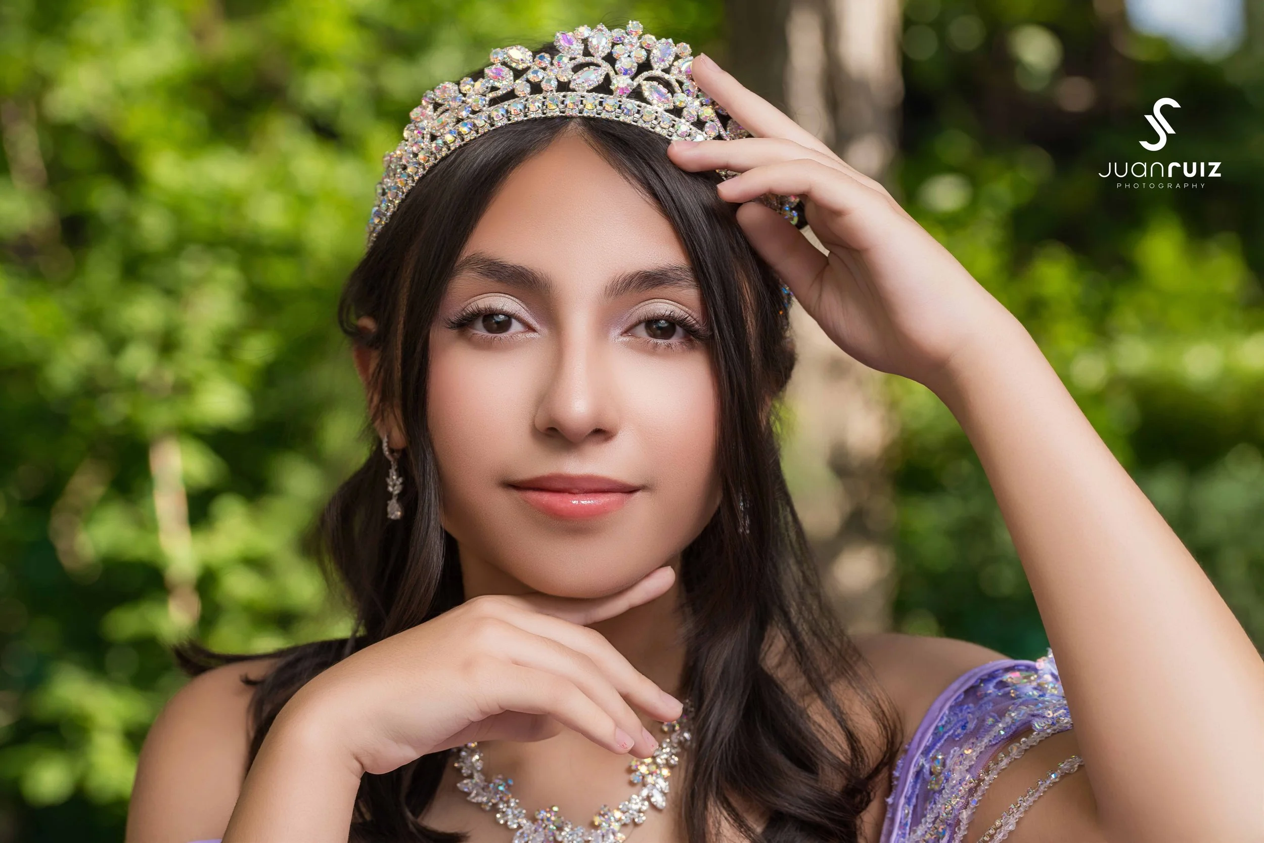 Young woman wearing a tiara and jewelry posing outdoors with a lush green background.
