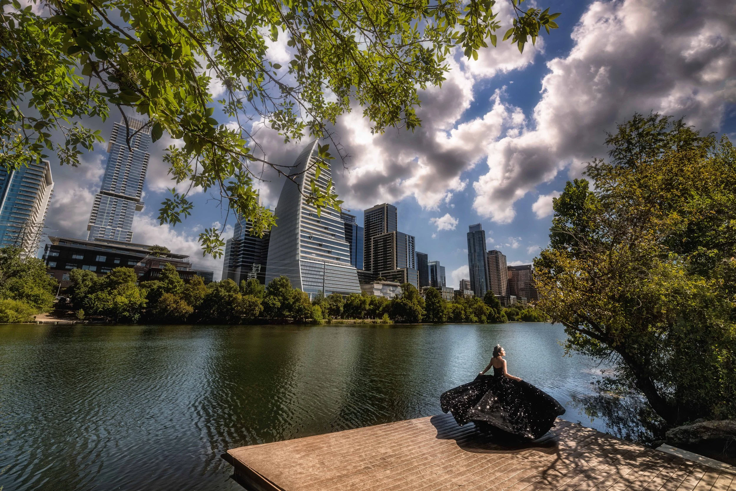 A woman in a long black dress sitting on a dock by the water, with a city skyline and tall skyscrapers in the background, under a partly cloudy sky.