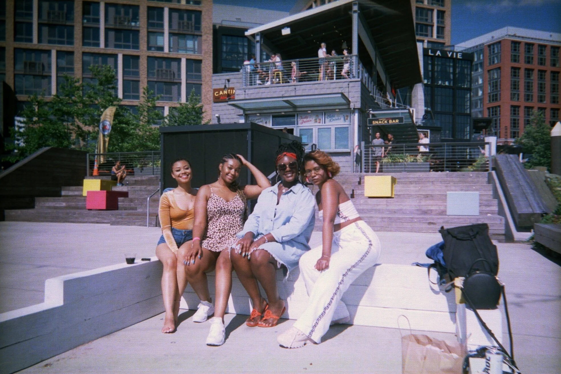 Four women sitting together on a low concrete bench in a sunny outdoor urban setting, with buildings and a multi-level patio with people in the background.