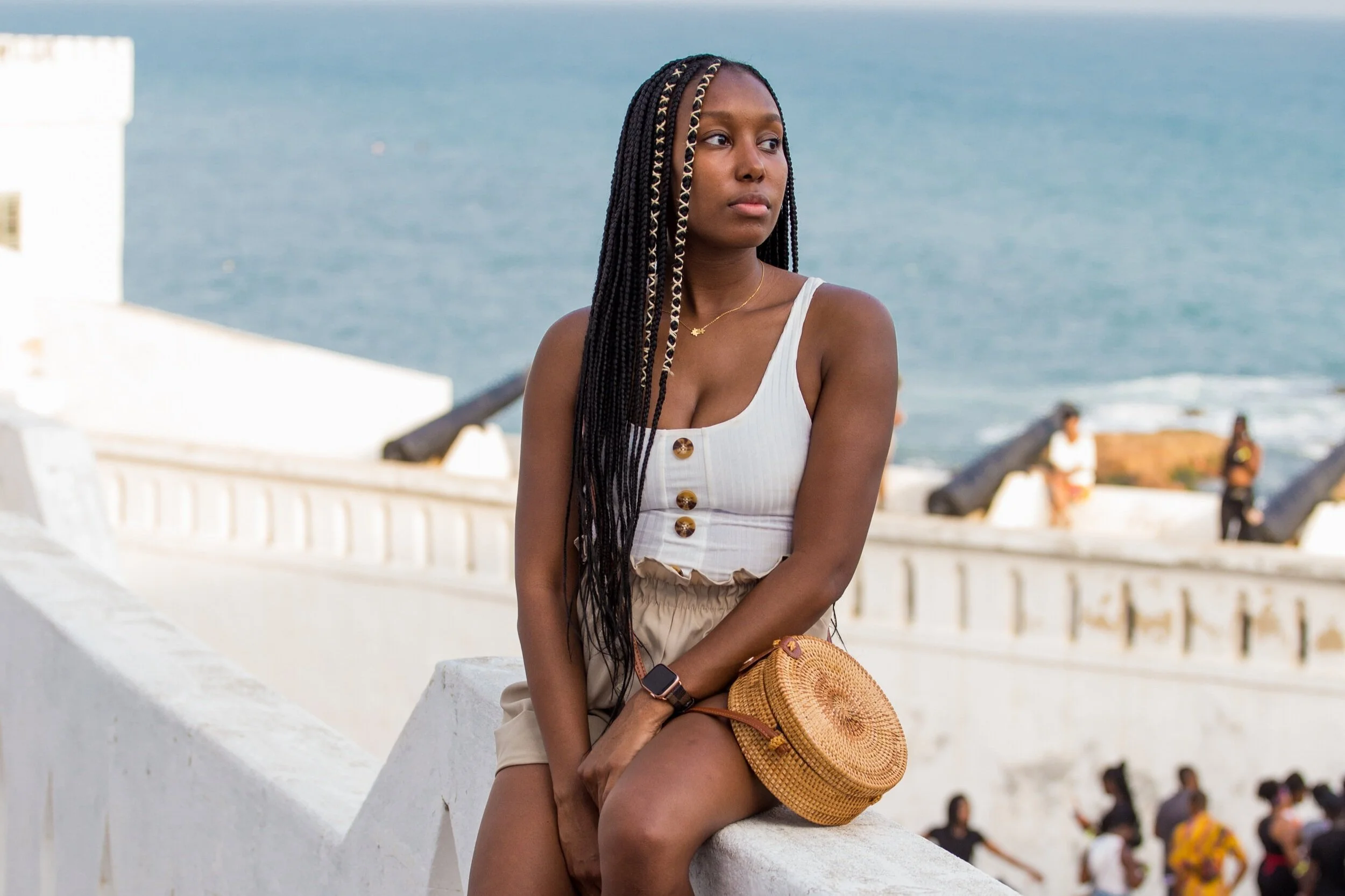 A young woman with long braided hair sitting on a white concrete barrier by the ocean, wearing a sleeveless white top with large buttons, beige shorts, a watch, and a round woven bag, with a blue sea and people in the background.