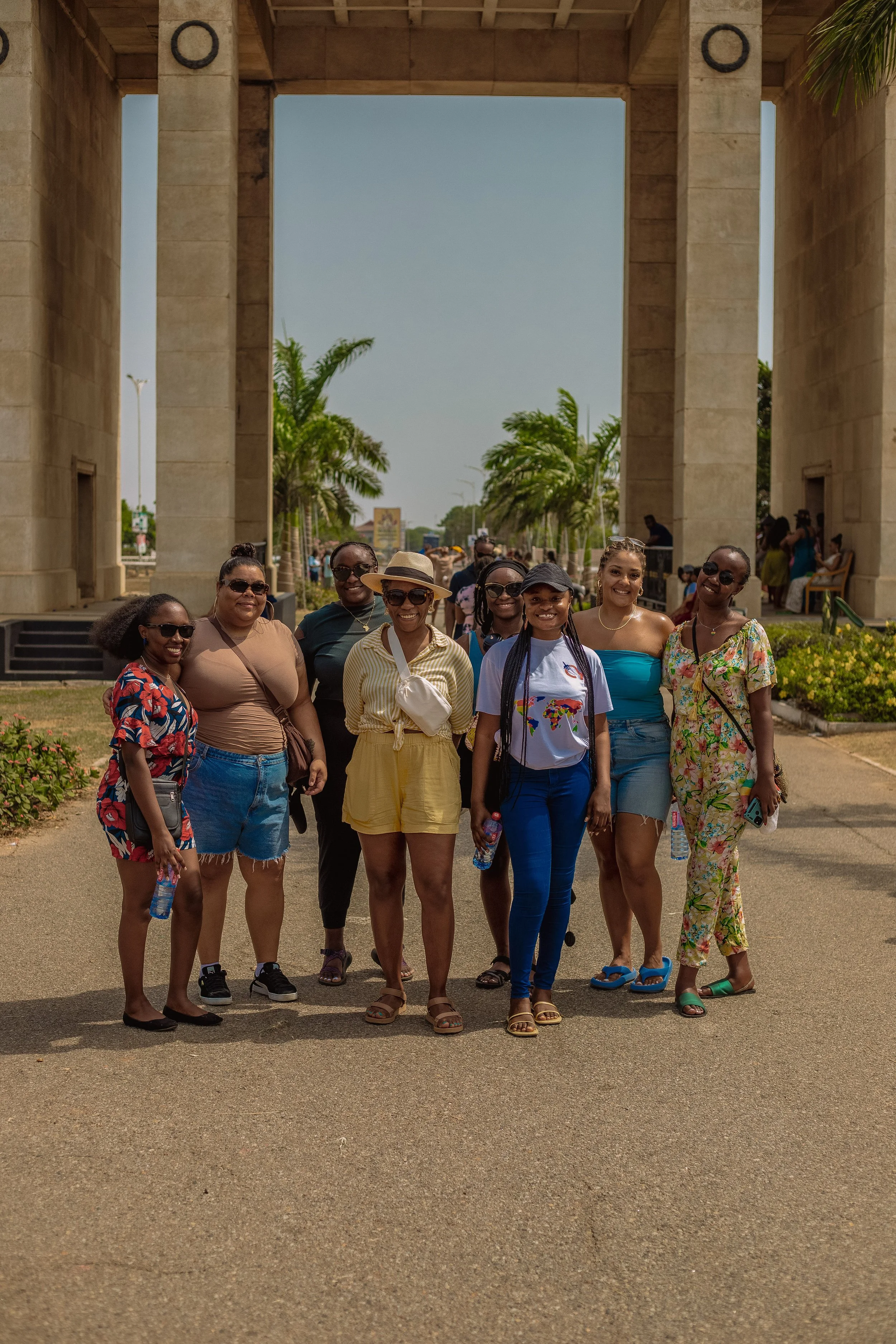 Group of women standing together outdoors in front of a large stone archway, smiling for a photo on a sunny day, some holding water bottles, with palm trees in the background.