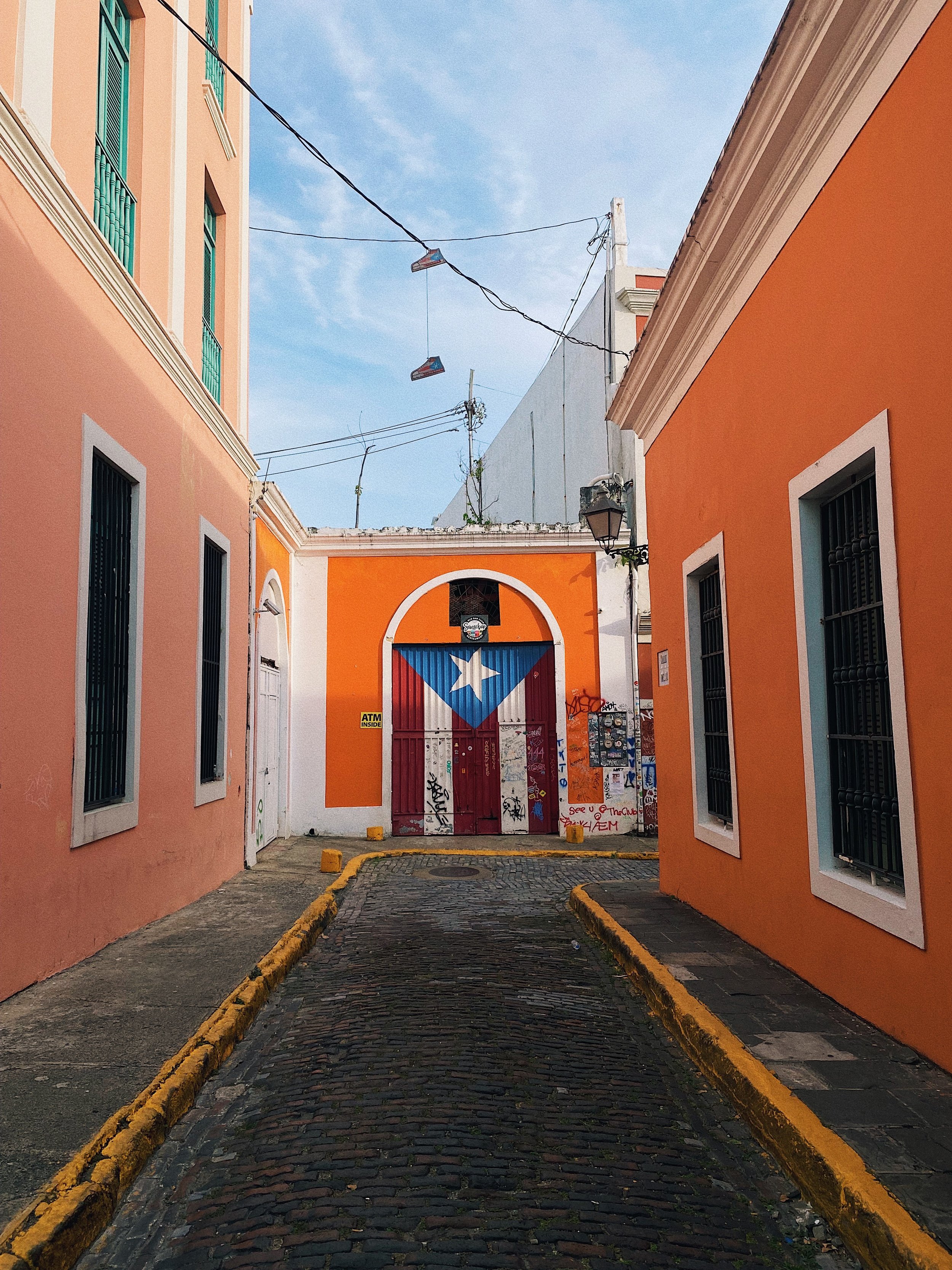 A narrow cobblestone street lined with colorful peach and orange buildings, with a Puerto Rican flag painted on a gate at the end of the street.