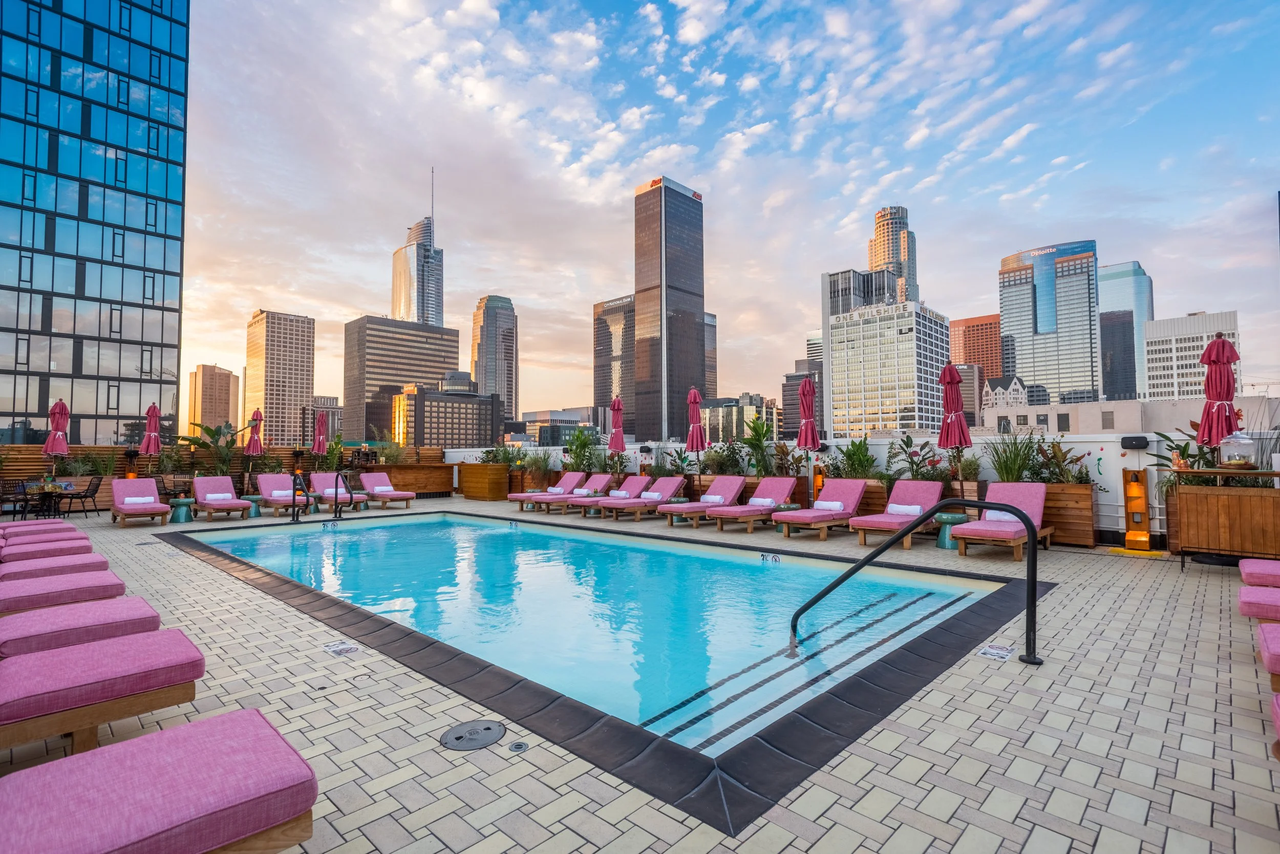 Rooftop swimming pool in Los Angeles with pink lounge chairs, umbrellas, and city skyline at sunset.