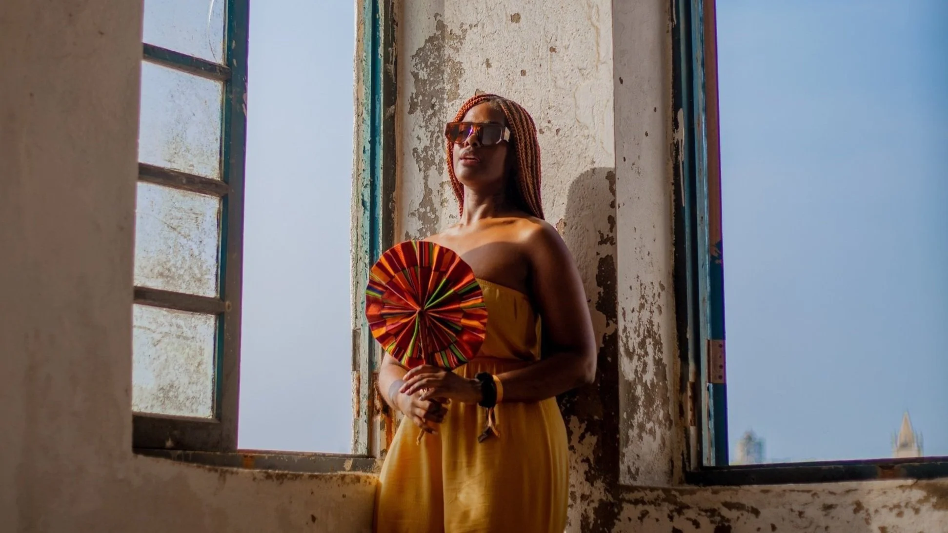 A woman with braided hair and large sunglasses standing by an open window in a weathered, textured wall, holding a colorful, circular fan.