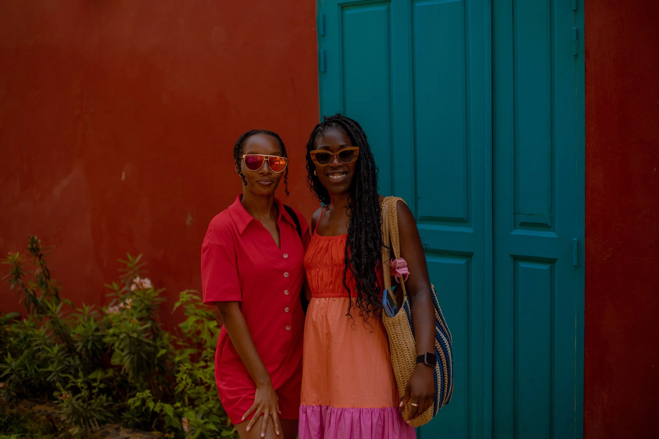 Two Black women wearing red and orange smile in front of a turquoise door on Goree Island in Dakar, Senegal