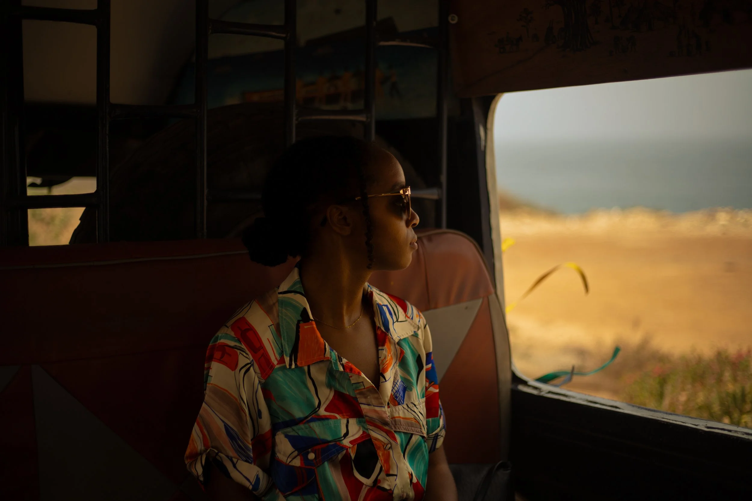 Black woman wearing pattern, multi-colored top with sunglasses staring out bus window in Dakar, Senegal