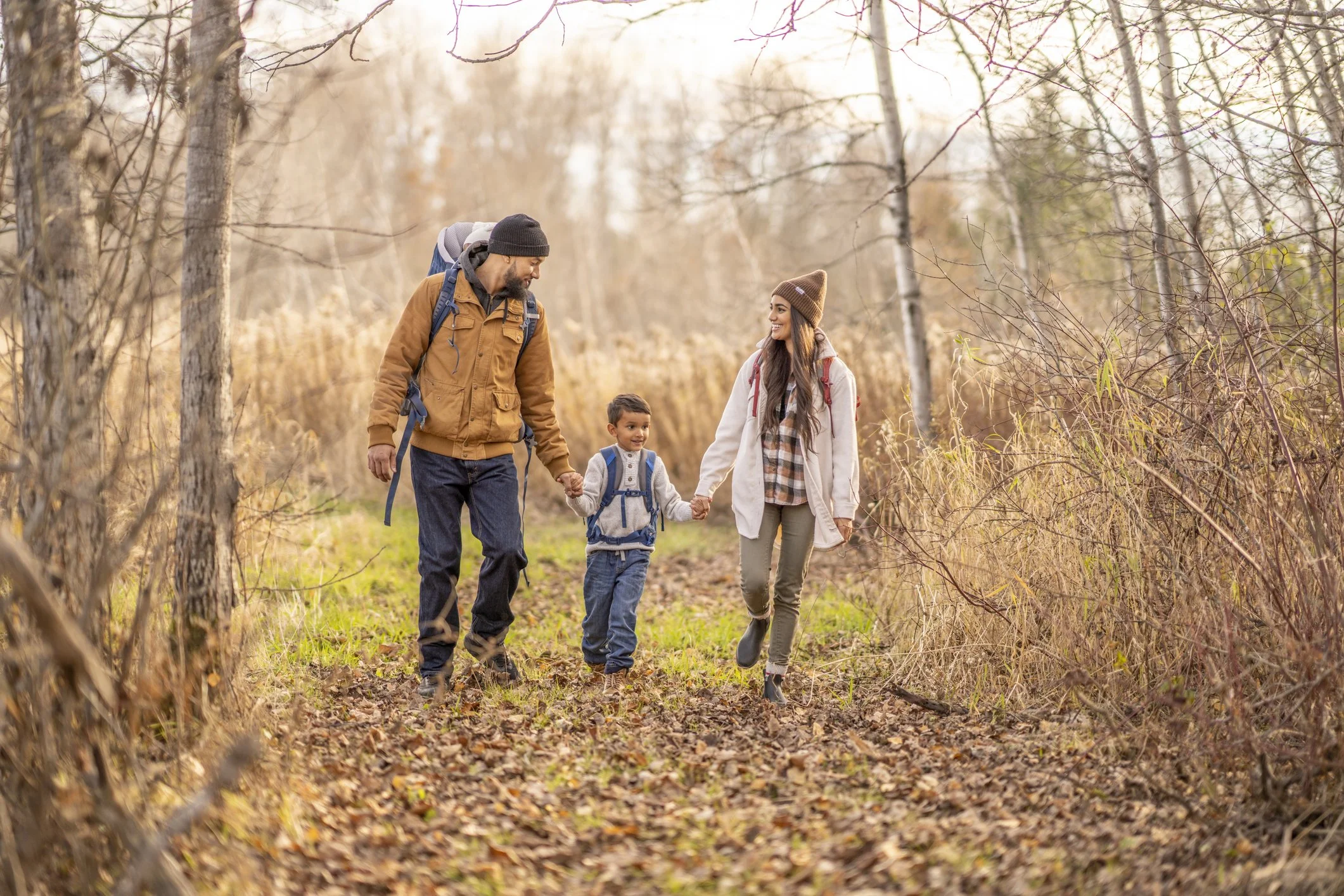 A family of three, a man, a woman, and a young boy, are walking along a dirt trail in a forest during autumn, holding hands and smiling.