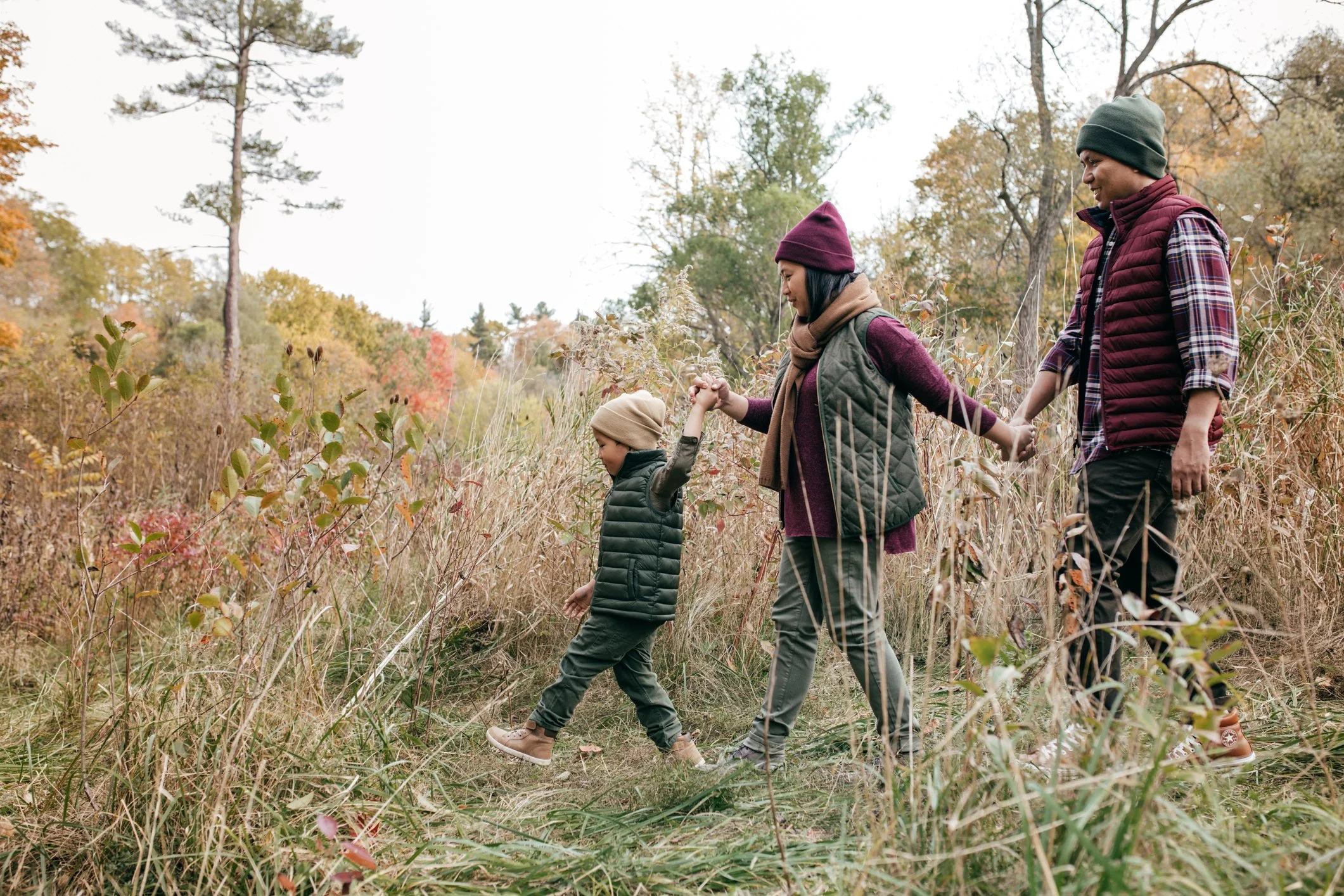 A family of three, wearing warm clothes and hats, holding hands and walking through a grassy, wooded area with autumn foliage.