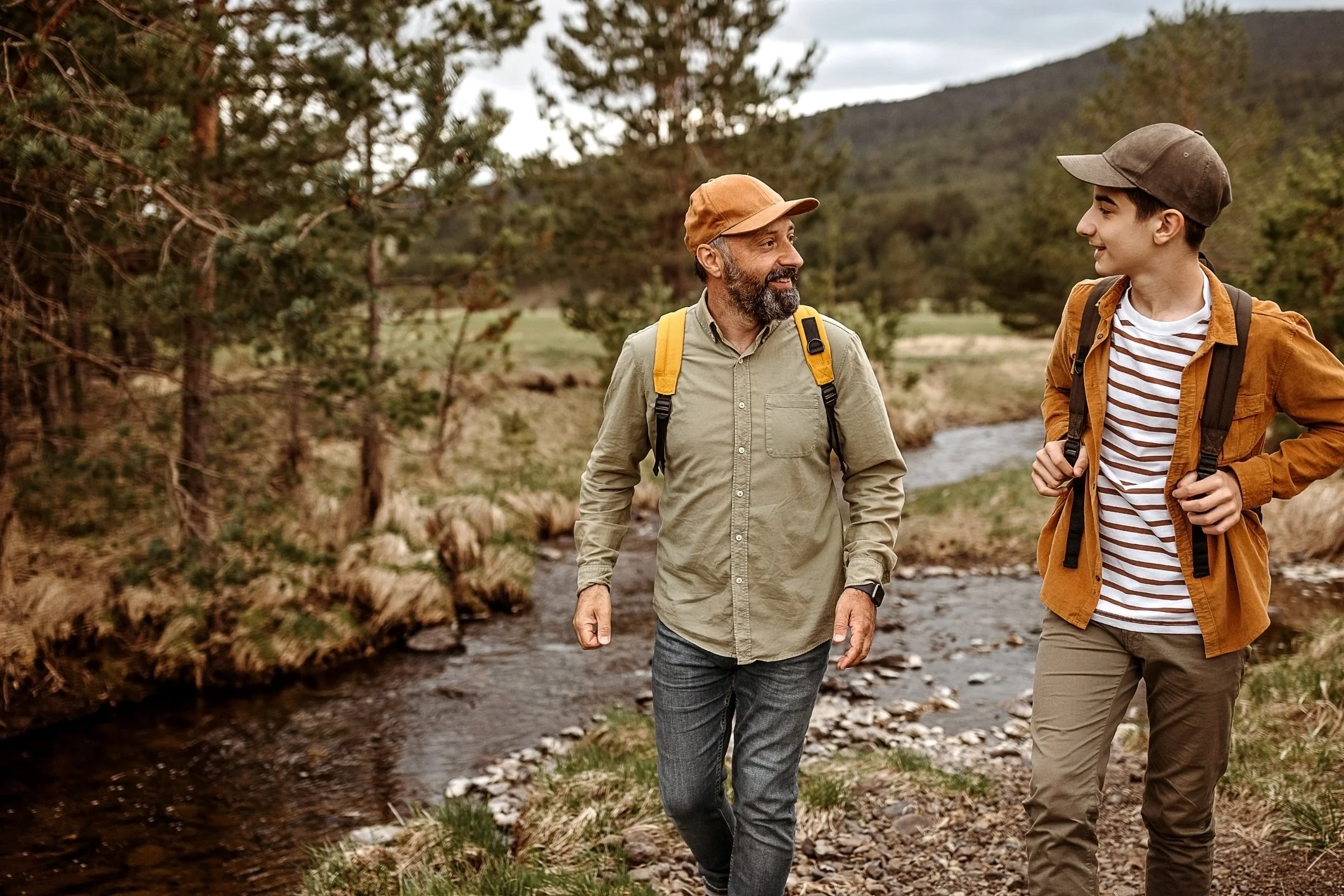 A man and a boy hiking in a forested area by a small stream, both wearing backpacks and outdoor clothing.
