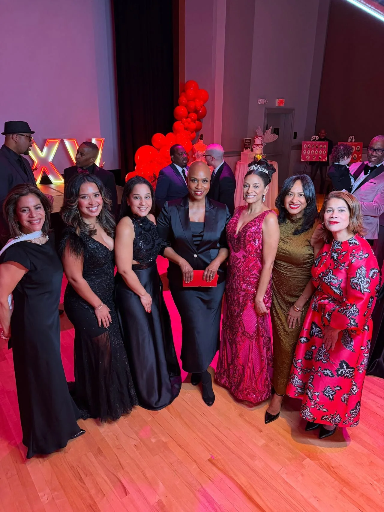 Group of elegantly dressed people at a black-tie event, smiling and posing for a photo on a red carpet. Background features red balloons, a neon sign, and evening party decorations.