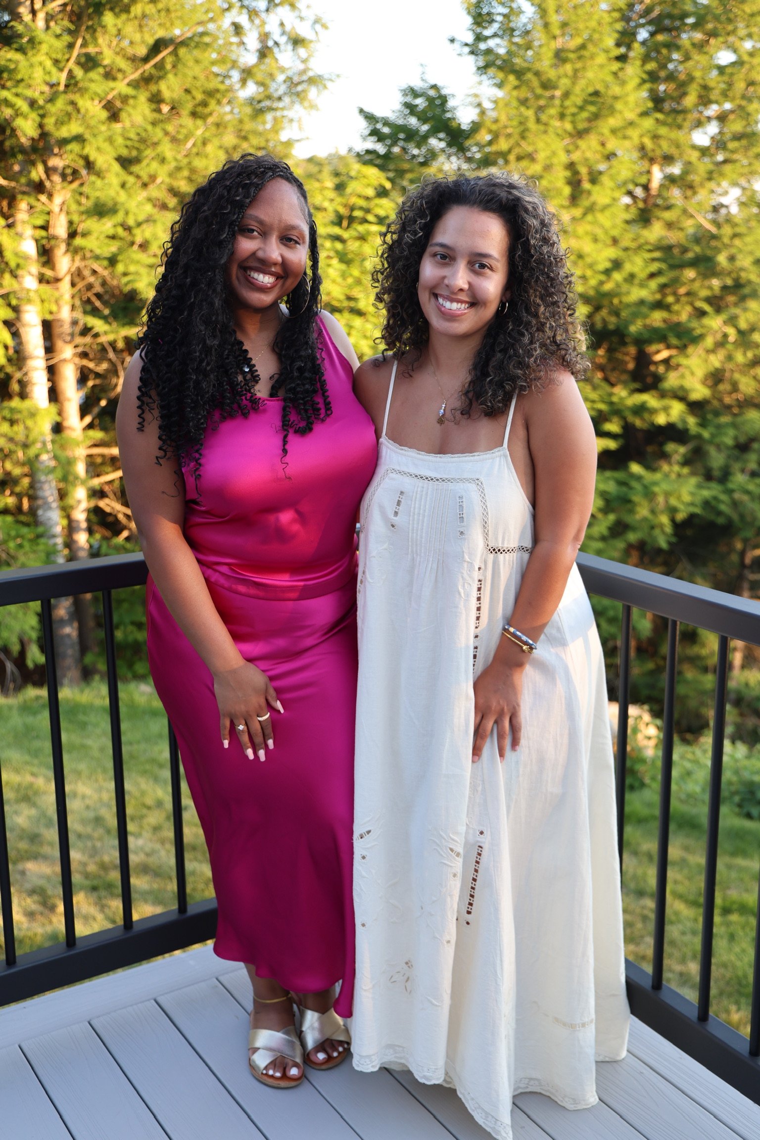 Two women standing outdoors on a wooden deck with a black railing, smiling at the camera with green trees in the background. The woman on the left is wearing a pink sleeveless dress and white sandals, and the woman on the right is wearing a white sleeveless dress and jewelry.