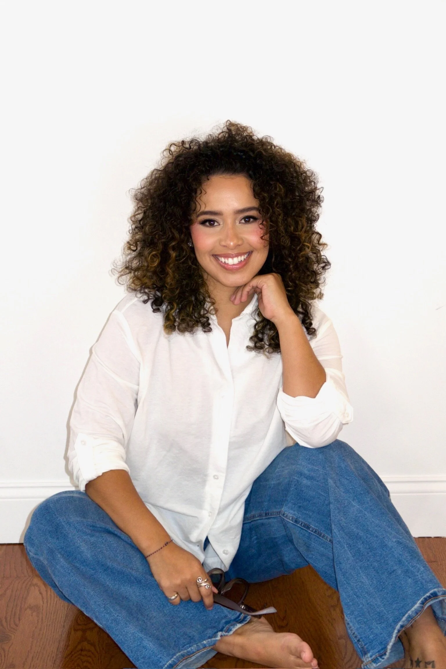 A woman with curly hair sitting on a wooden floor against a white wall, wearing a white shirt and blue jeans, smiling and touching her chin with her right hand.