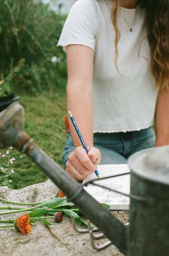 Person in a garden, drawing in a notebook, surrounded by gardening tools and flowers.