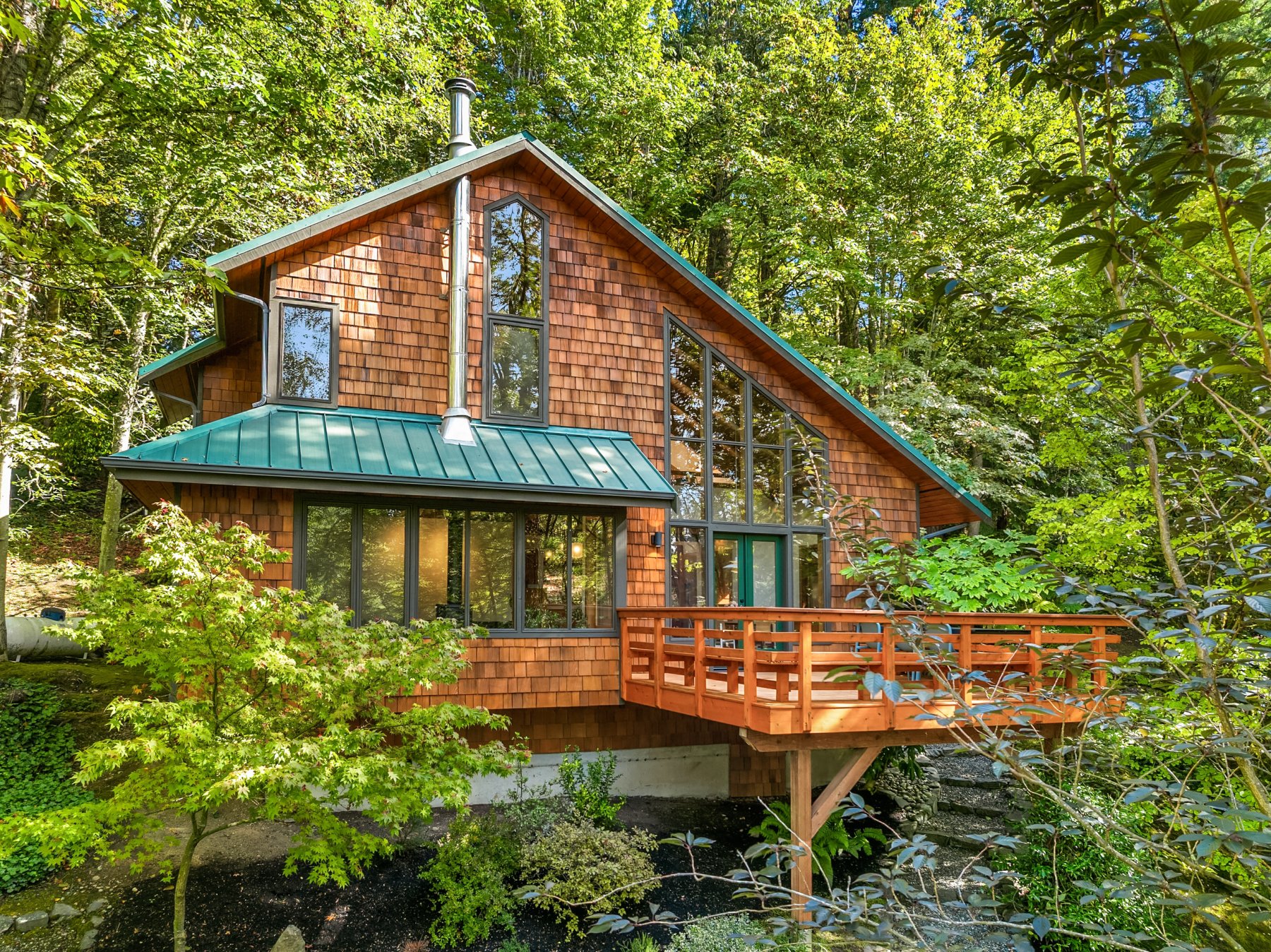 Wooden house with large glass windows surrounded by green trees and shrubbery.