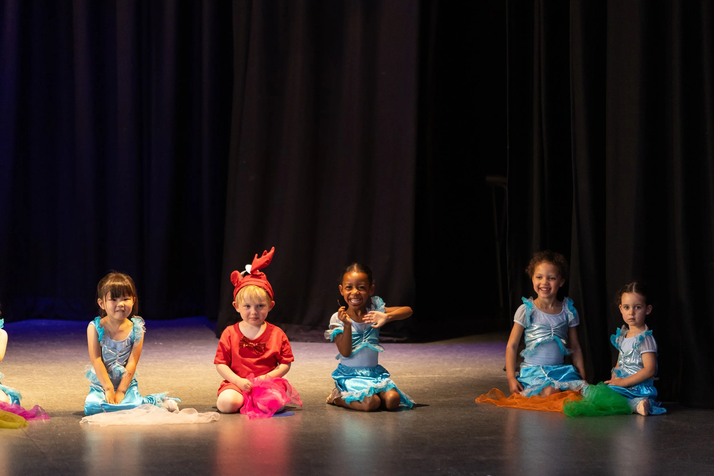Five young girls dressed in colorful costumes sitting on a stage with black curtains in the background.