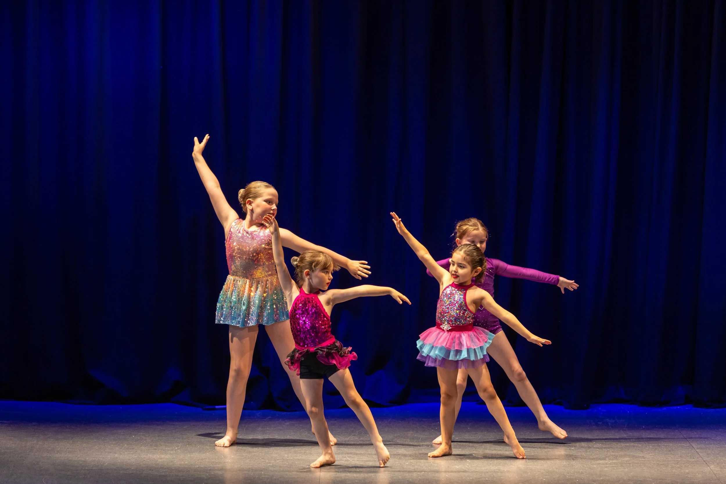 Four young girls in colorful costumes performing a dance on stage with blue curtain backdrop.