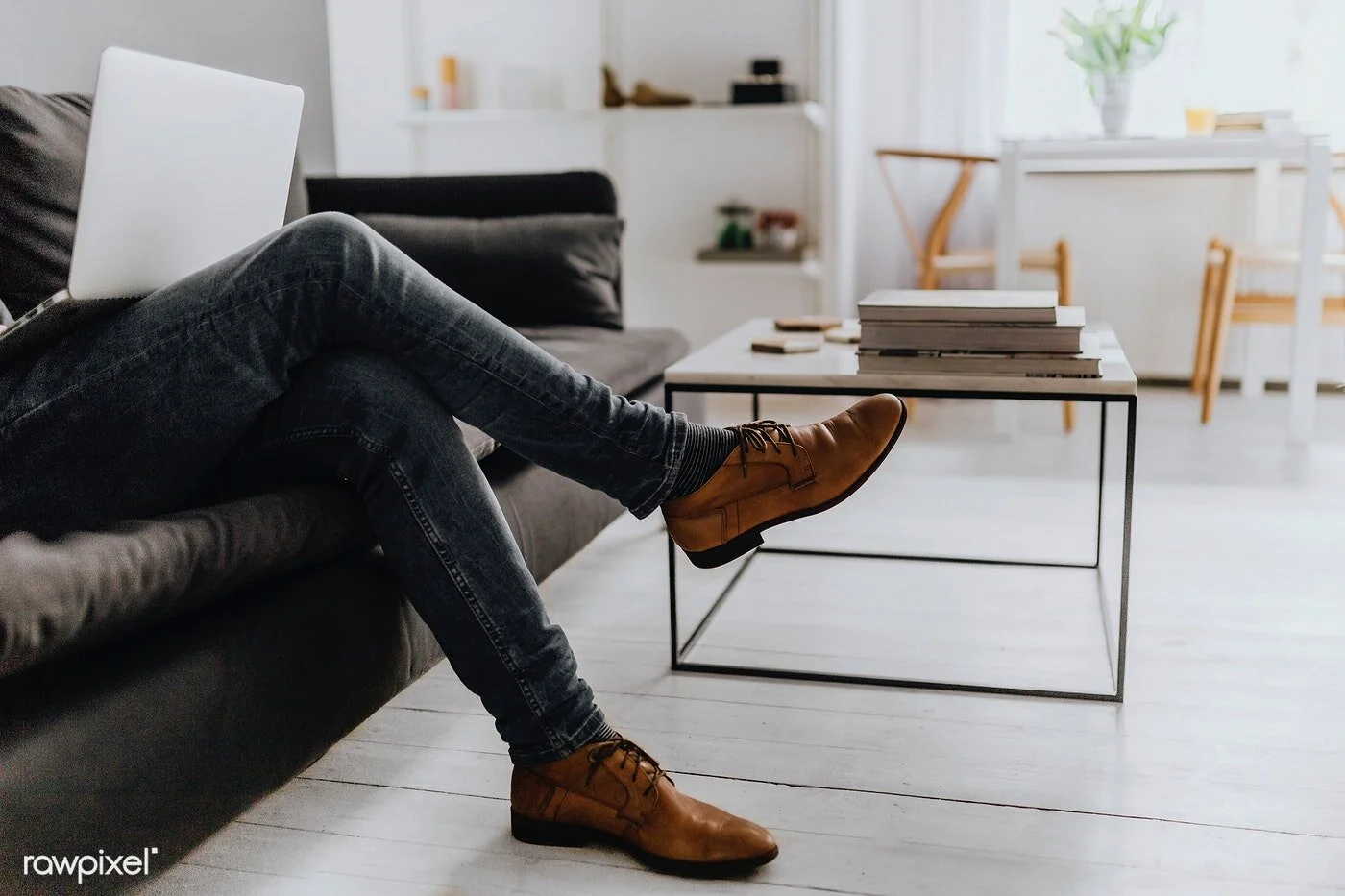 A man sitting on a gray sofa in a bright, modern living room with legs crossed, wearing dark jeans and brown leather shoes, using a laptop.