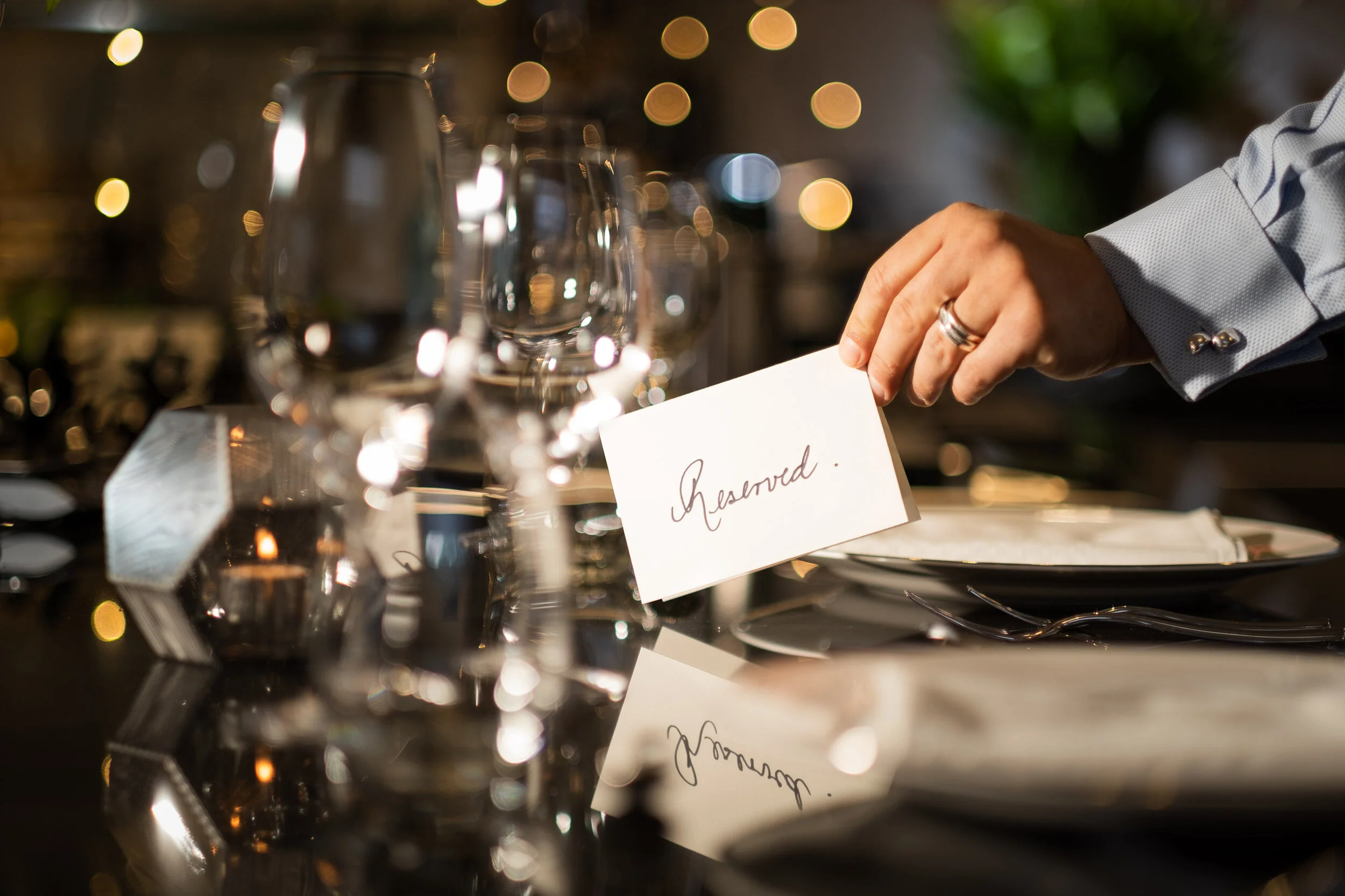 A table setting at a restaurant with wine glasses, silverware, and a reserved sign, with blurred warm lights in the background.