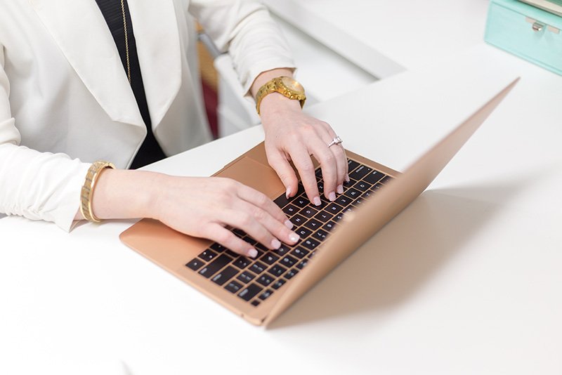 Woman typing on a gold-colored laptop with a white desk and pastel blue storage box in the background.