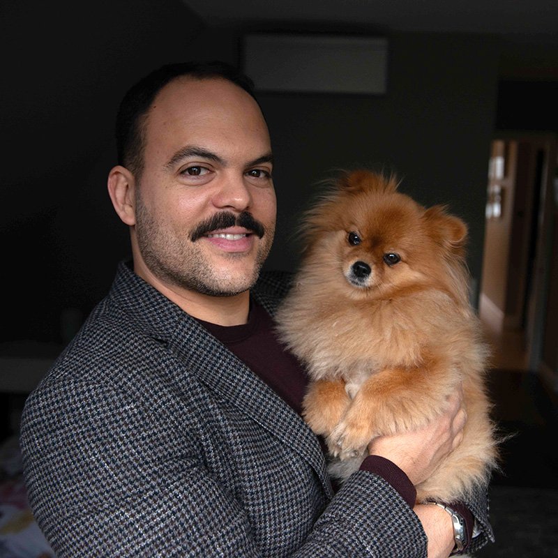 Victor Arraez holding a small fluffy Pomeranian dog indoors.