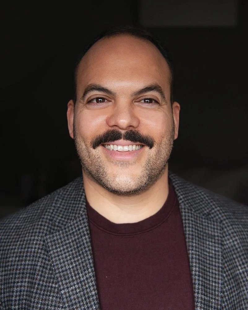 Victor Arraez wearing a checkered blazer and a maroon shirt against a dark background.