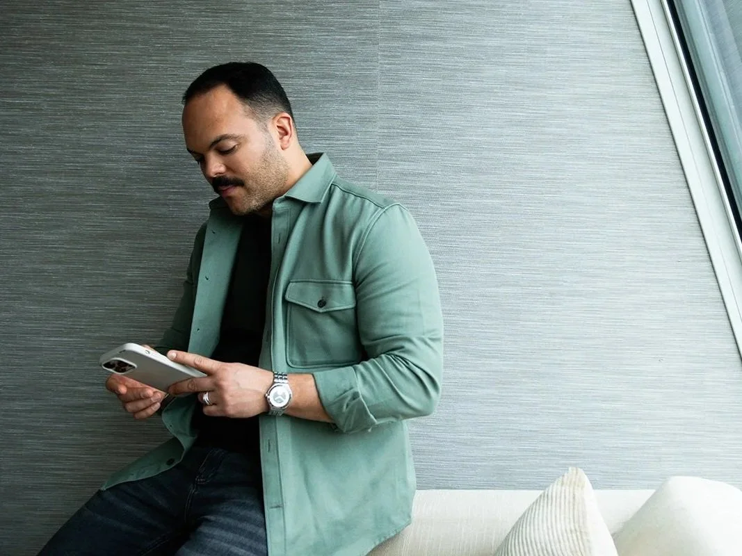 Victor Arraez wearing a green jacket, black shirt, and a watch, sitting on a white couch and looking at his smartphone.