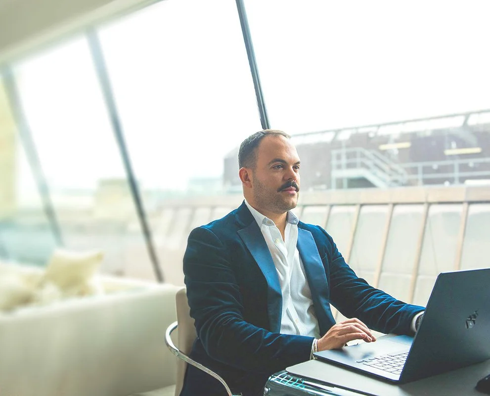 Victor Arraez  in a dark blue blazer and white shirt working on a laptop in a modern office with large windows and a cityscape view.