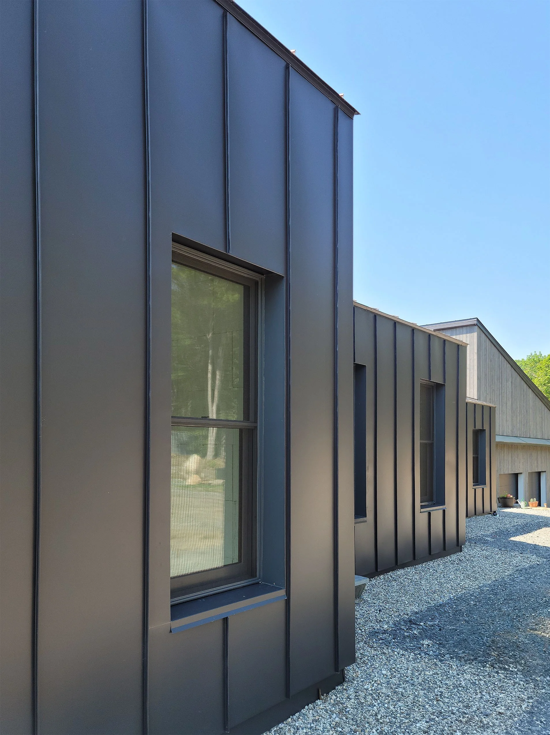 Side view of a modern building with dark gray or black metal siding and large windows, surrounded by a gravel pathway.