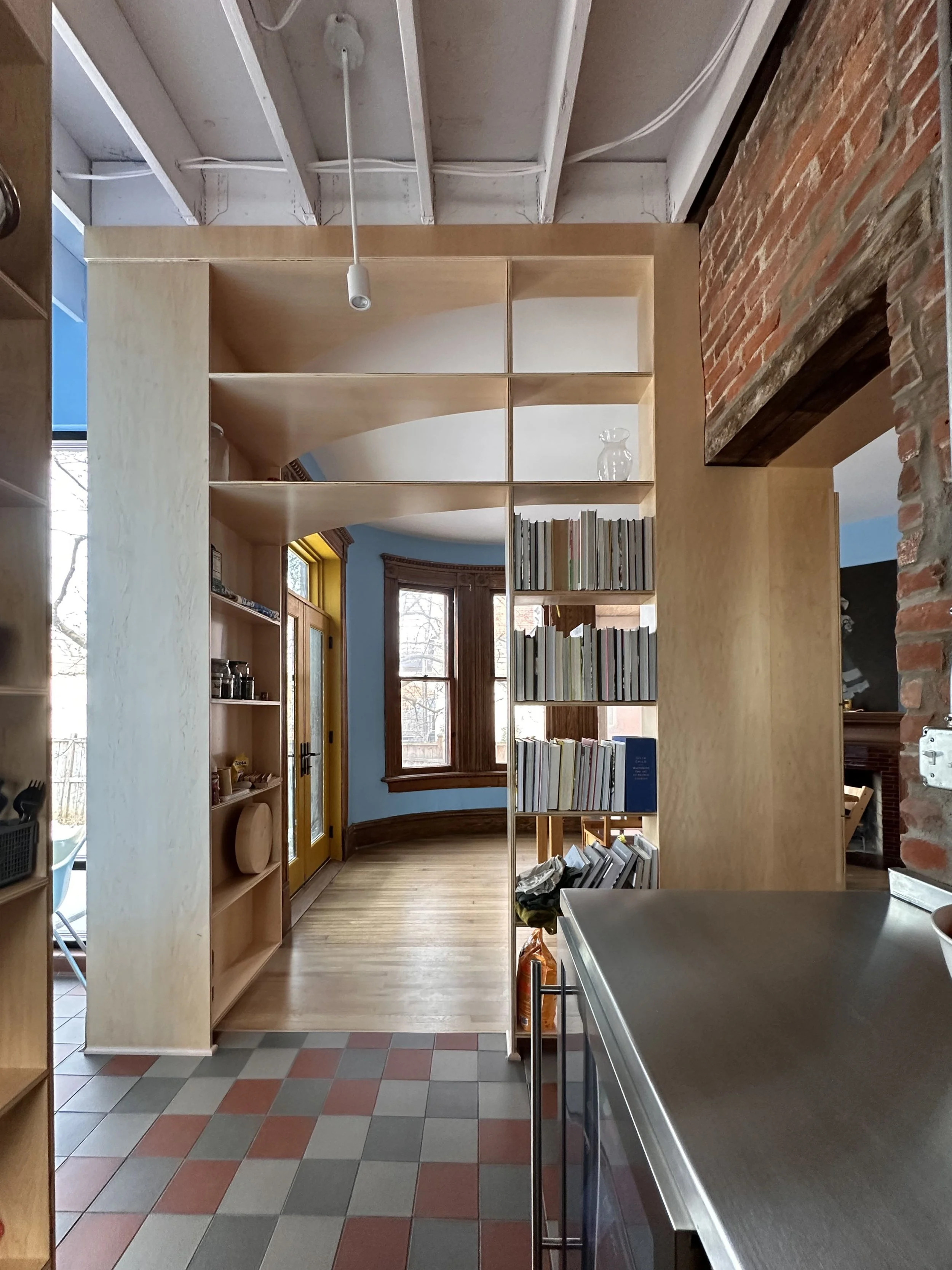 Interior of a cozy home with a wooden bookshelf acting as a room divider, large blue walls, windows with wooden trim, and decorative brickwork. Metal countertop in the foreground.