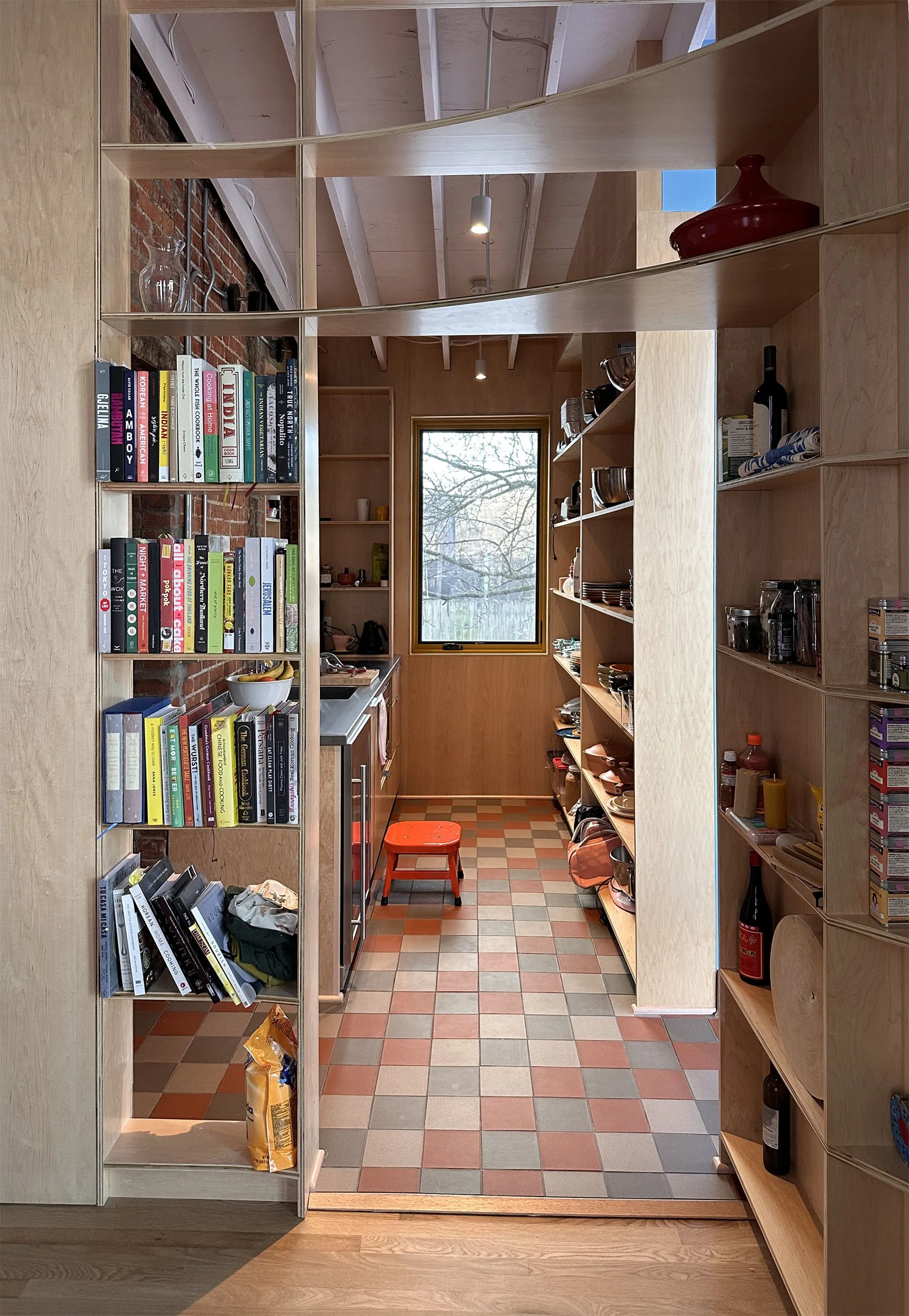 View through a doorway into a kitchen with wooden shelves, a window showing outdoor trees, and a tiled floor with red and beige squares. Shelves hold dishes, bottles, and kitchen items, and there is a red stool in the kitchen.