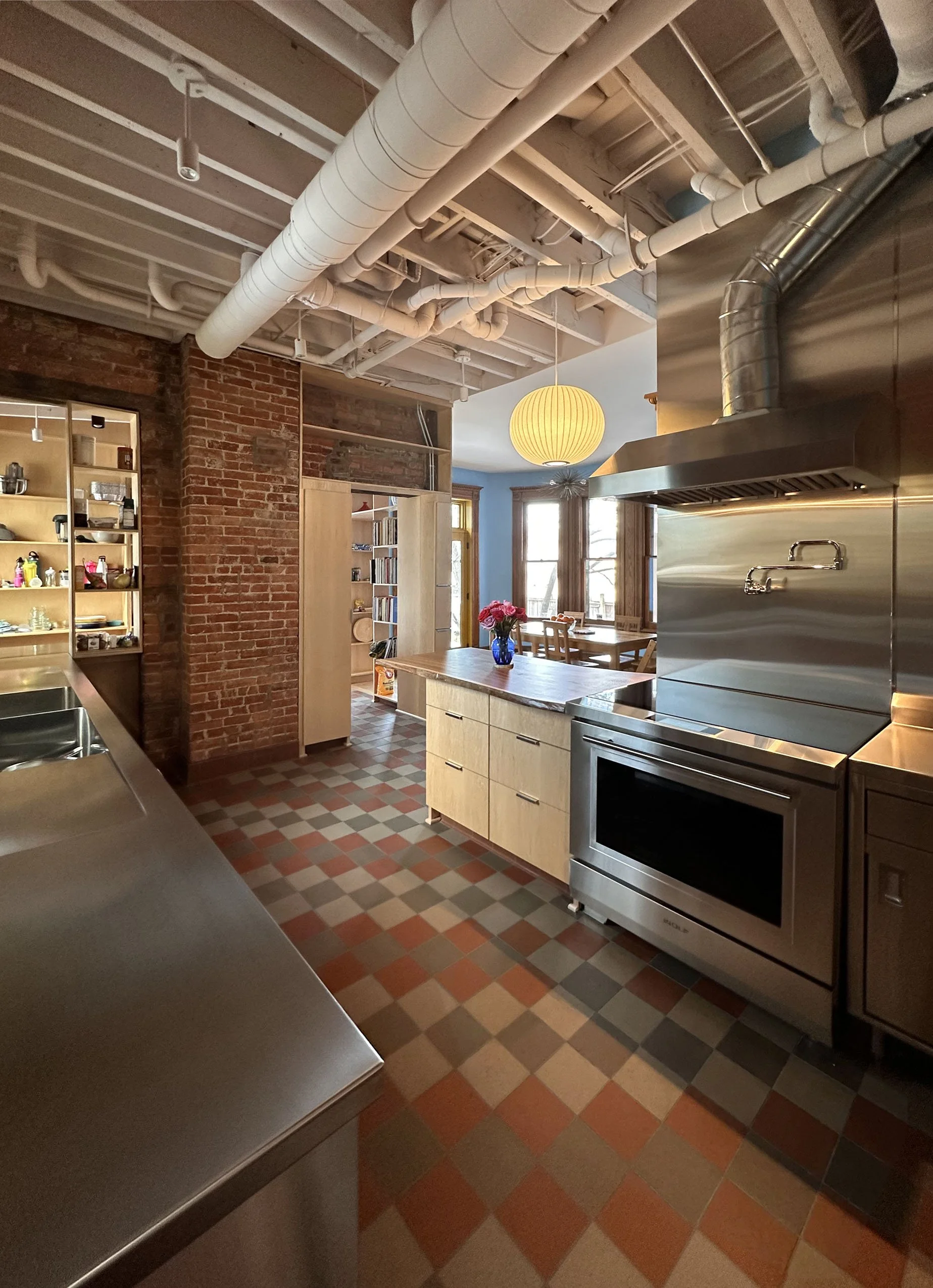 A modern kitchen with stainless steel appliances, wooden cabinets, a brick wall, and a round yellow light fixture hanging from the ceiling. There is a table with flowers and windows in the background.