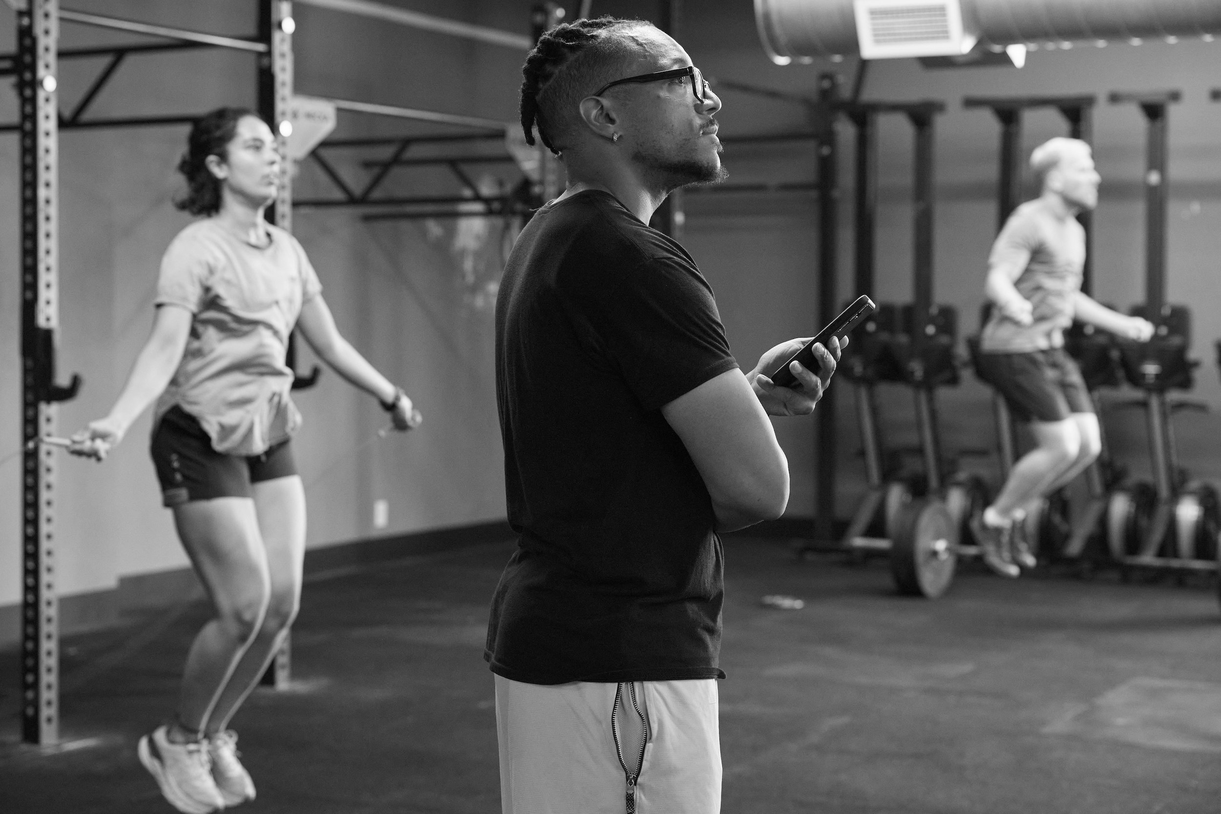 A man with glasses and styled hair looking at his phone in a gym. In the background, two women are jumping rope, one wearing a short-sleeve shirt and shorts, and the other in athletic wear. The gym has power racks and exercise equipment.