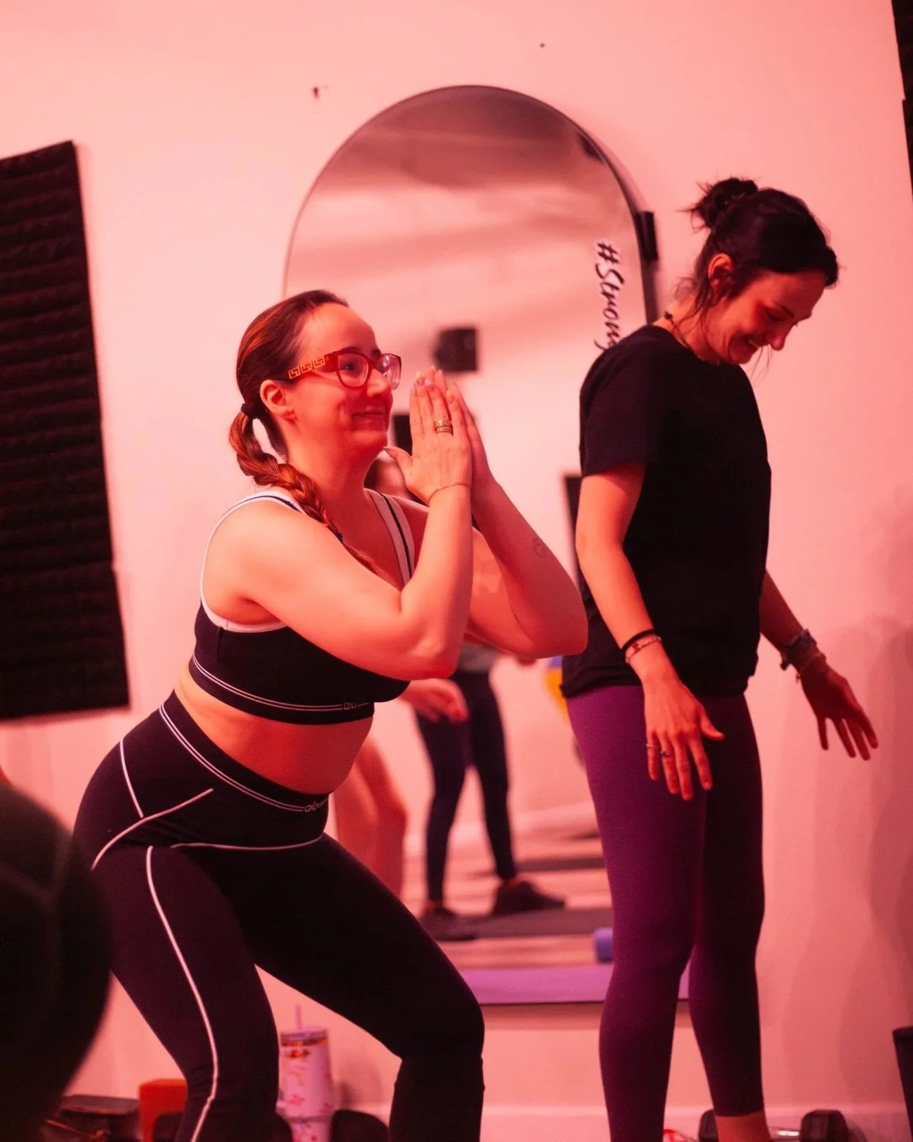 Two women practicing yoga in a fitness class, one in a black sports bra and leggings with white lines, and the other in a black shirt and purple leggings, both smiling and standing on yoga mats.